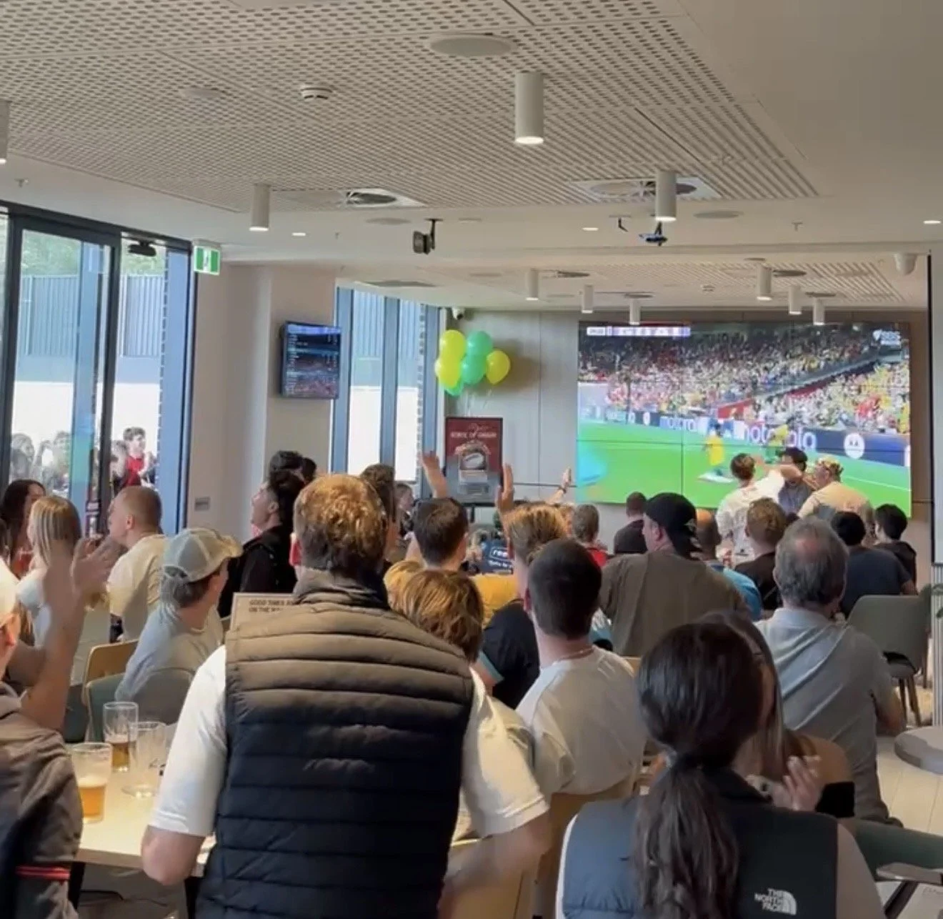 People watching a sports event on a large screen in an indoor viewing area with balloons and large windows.