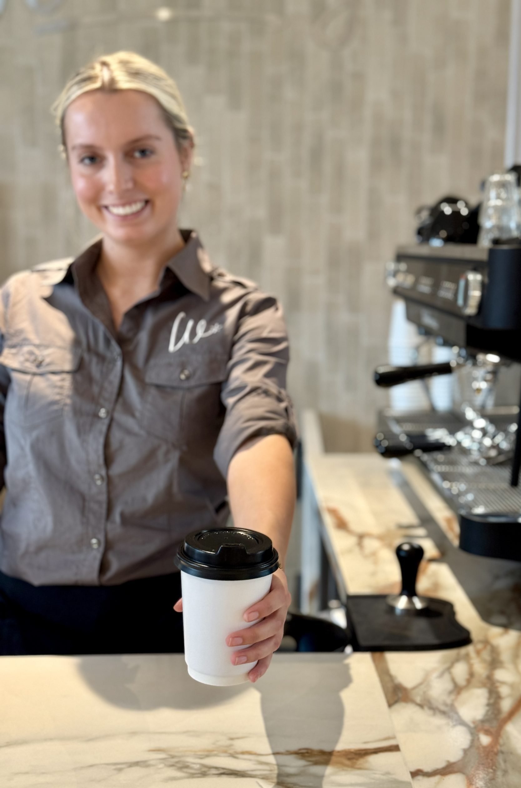 A smiling woman with blonde hair holding a white disposable coffee cup with a black lid inside a coffee shop or cafe.