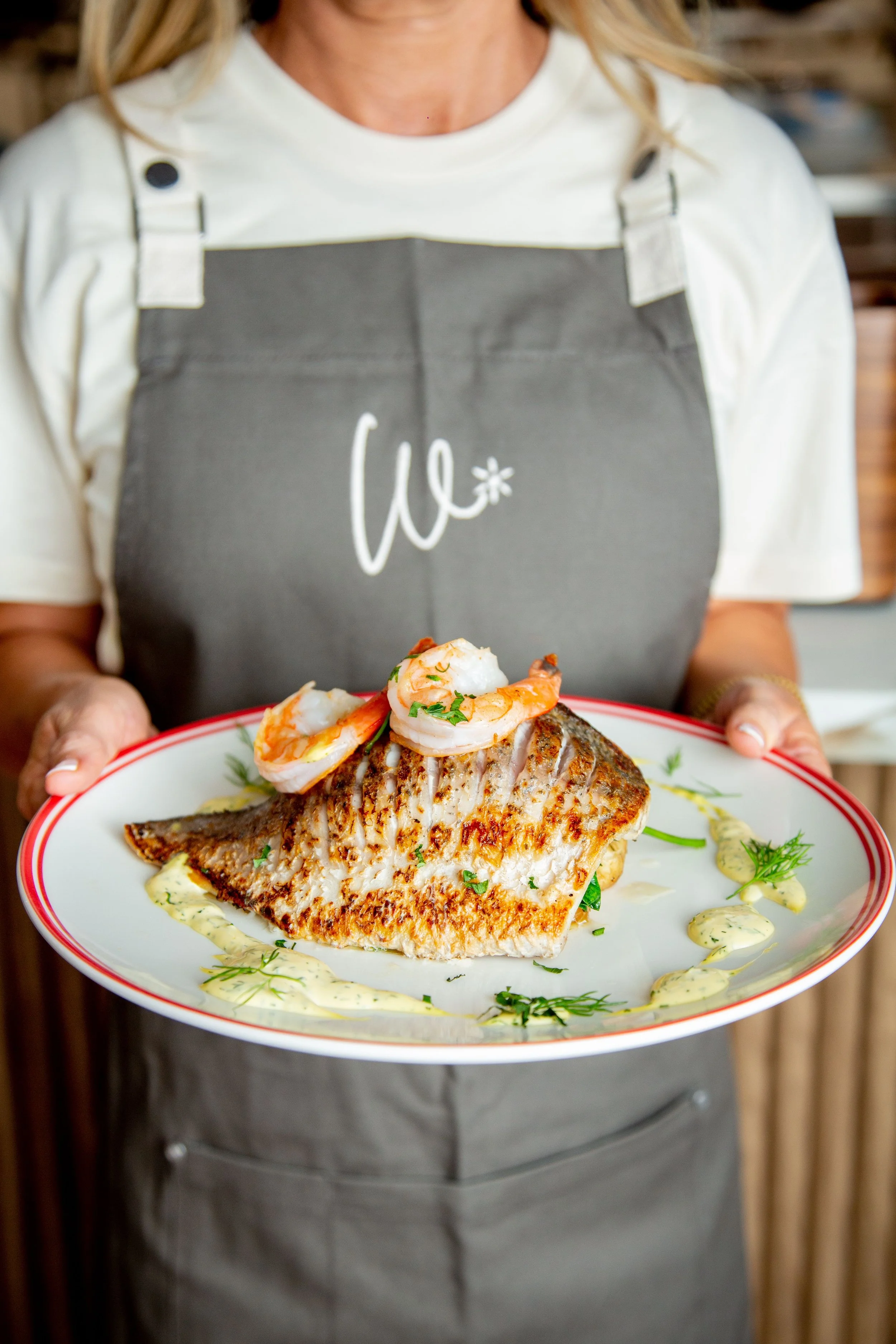 A person wearing a white t-shirt and a gray apron with 'WICI' written on it is holding a plate with a cooked fish fillet topped with cooked shrimp and garnished with herbs, served with a creamy sauce.
