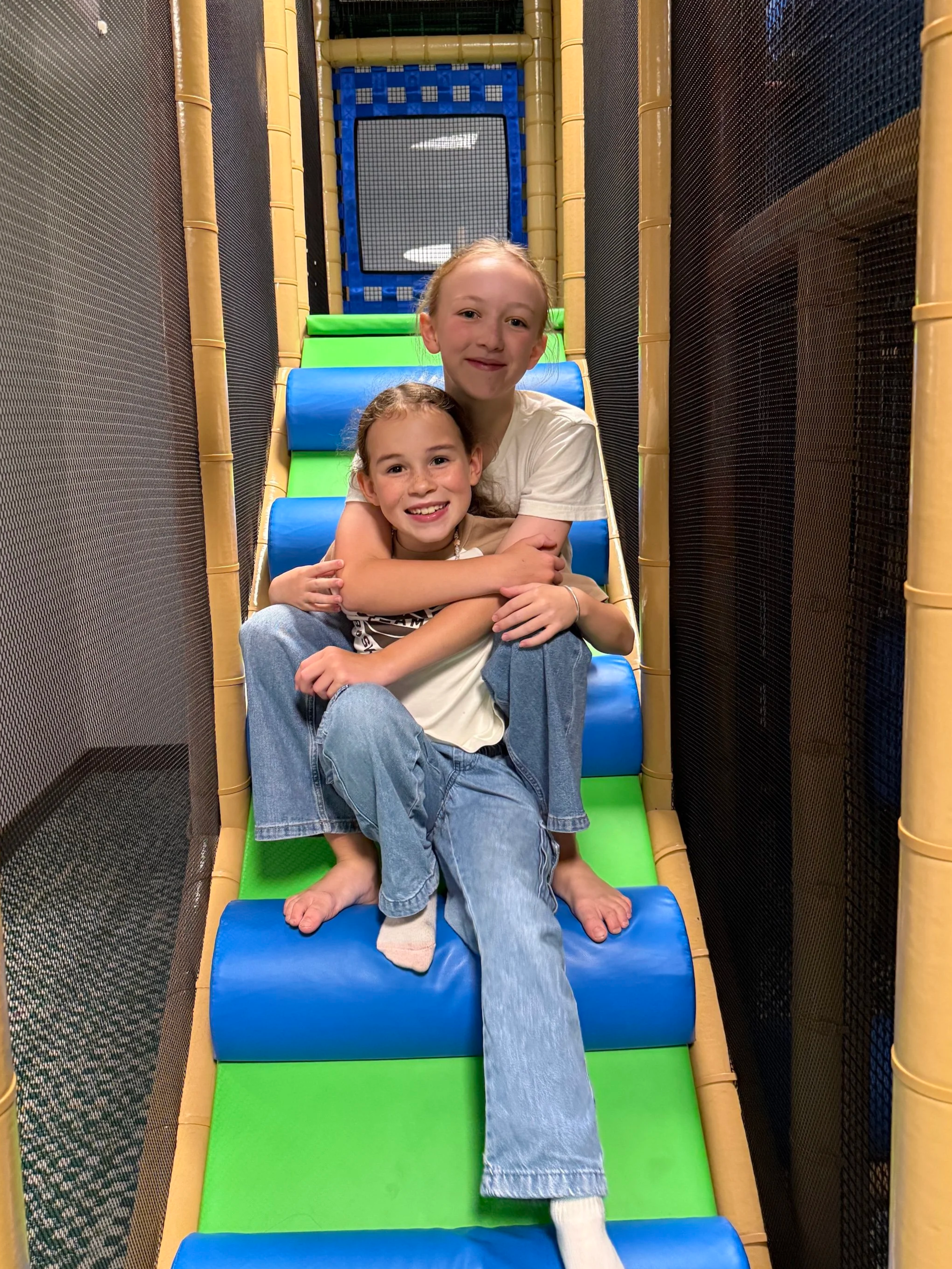 Two smiling young girls sitting on an indoor climbing slide, with padded blue, green, and yellow sections, surrounded by black safety netting.