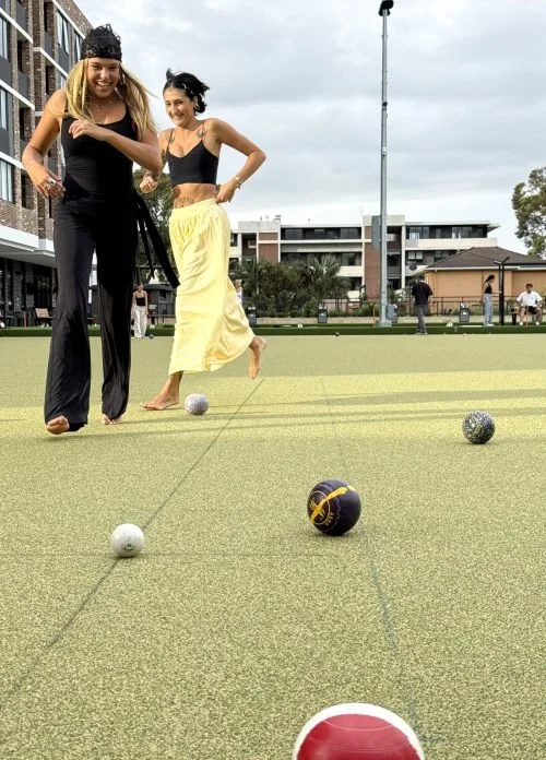 Two women playing lawn bowls on an outdoor green, with several bowls and a jack in play, surrounded by other people and buildings.
