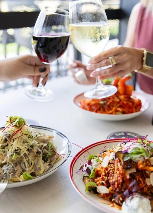 Two people clinking glasses of red and white wine at a table with pasta dishes and a plate of spaghetti with tomato sauce in the background.