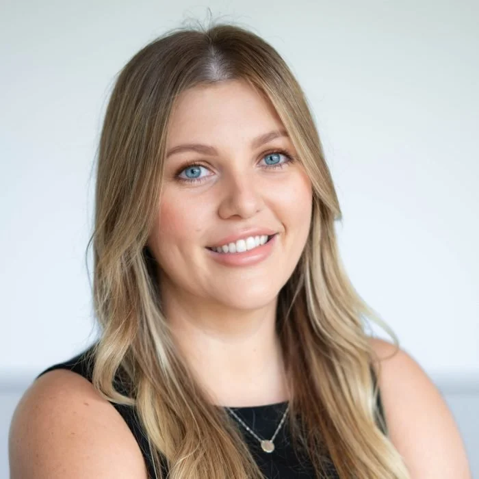 A young woman with long blonde hair and blue eyes smiling at the camera, wearing a sleeveless black top and a necklace, with a blurred light background.