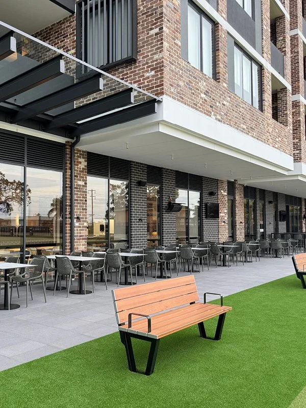 Outdoor seating area with a row of black tables and chairs, green artificial turf, and a brick building with large windows and a balcony.