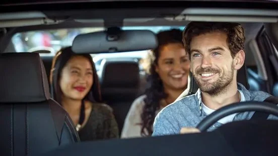 A man driving a car with two women sitting in the backseat, all smiling and enjoying the ride.