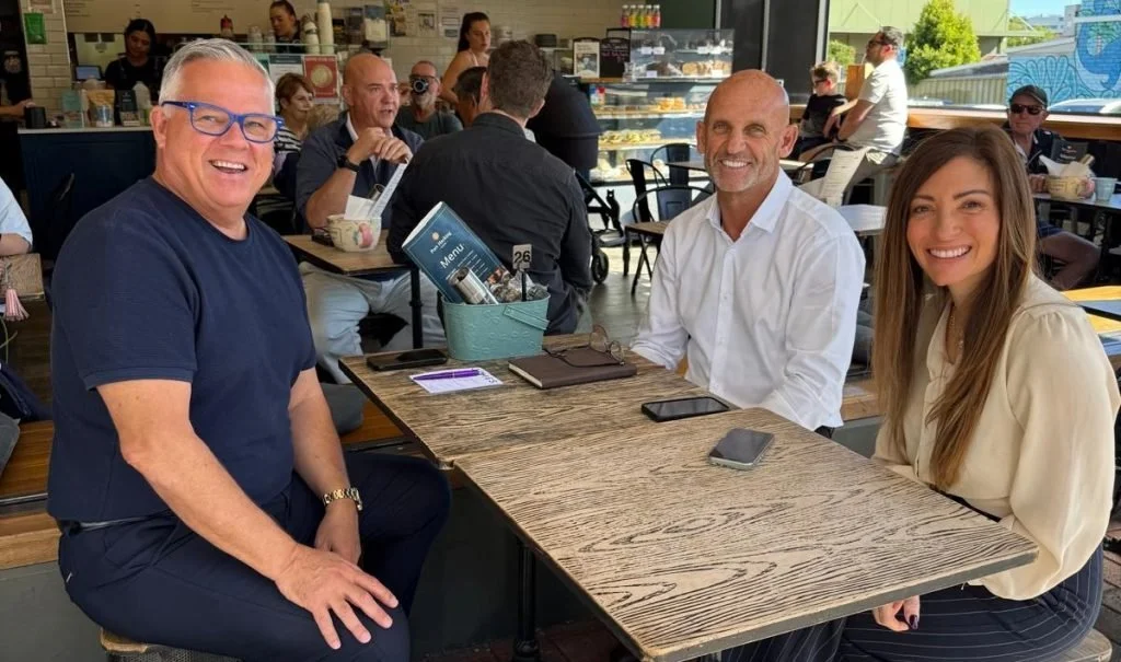 Three people sitting at a wooden table in a restaurant, smiling at the camera. The person on the left is a man with gray hair and glasses wearing a navy blue shirt. The person in the middle is a man with a bald head and a white shirt. The person on the right is a woman with long brown hair wearing a beige blouse. There are other diners and a view of outdoors in the background.
