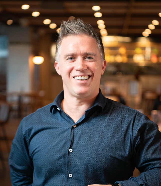 A smiling man with short, spiked gray hair wearing a dark blue button-up shirt in a warmly lit restaurant or cafe.