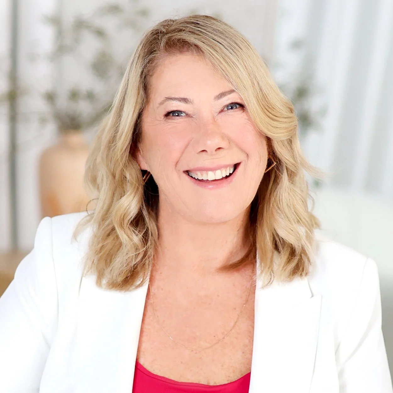 A smiling middle-aged woman with blonde wavy hair, wearing a white blazer and a pink top, sitting indoors with a blurred background of plants and neutral decor.