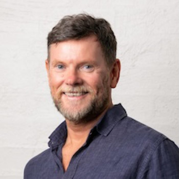 A smiling middle-aged man with short brown hair and beard, wearing a navy blue shirt, standing against a light-colored wall.