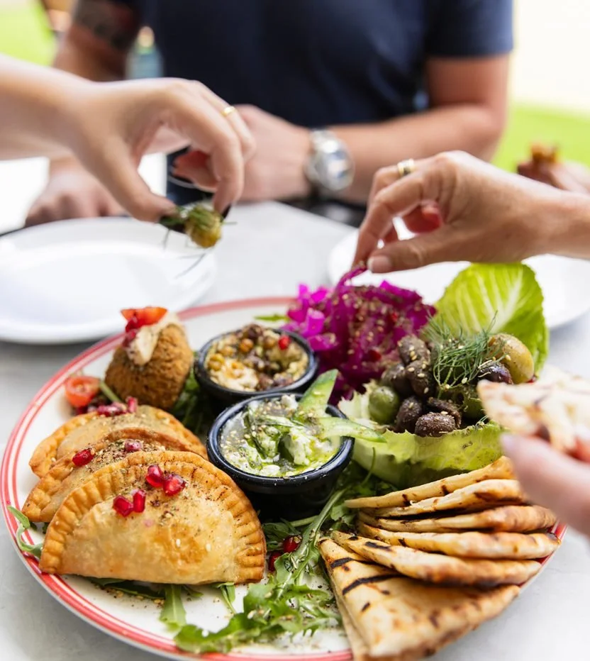 A plate with an assortment of Middle Eastern dishes including grilled flatbread, stuffed pastries, hummus, tabbouleh, and fresh vegetables, with people reaching for food in the background.
