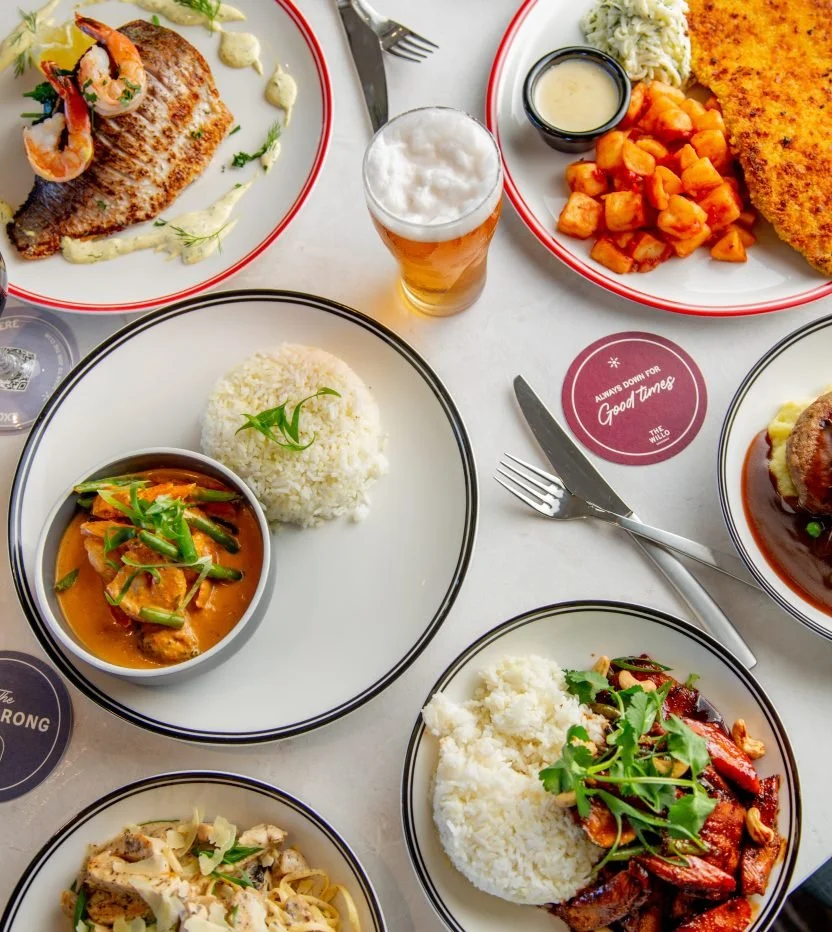 Assorted plates of food including grilled fish with shrimp, stew with rice, fried fish with sweet potatoes, chicken with mashed potatoes, and a bowl of pasta, accompanied by a glass of beer on a table.