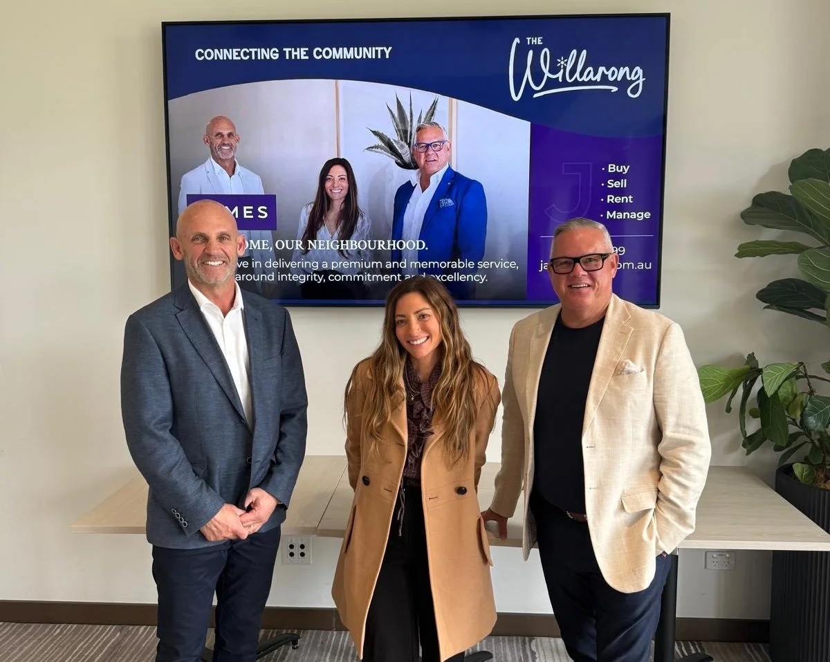 Three people standing in front of a large screen displaying a presentation slide. The screen shows a group photo of three individuals and the words "Connecting the Community" and "The Willarang". The people in front are smiling and dressed in business casual attire; two men in blazers and a woman in a tan coat.