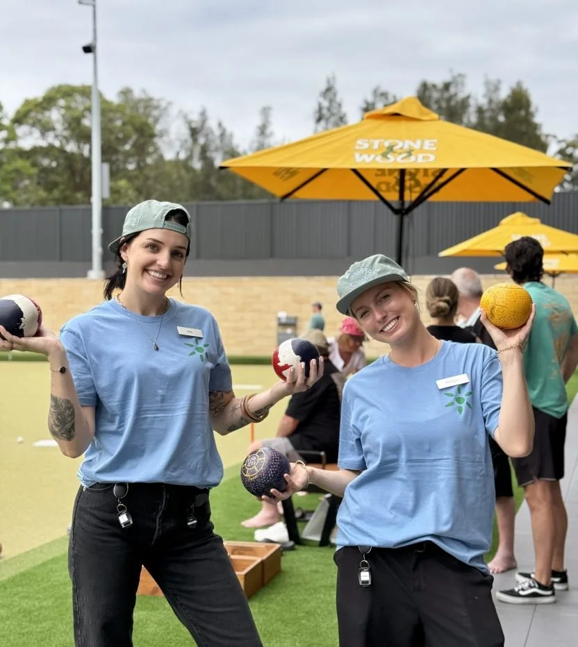 Two smiling women wearing blue shirts and light gray caps holding colorful balls at an outdoor event with yellow umbrellas and people in the background.