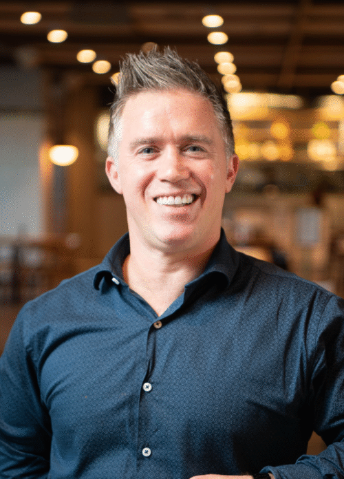A smiling man with short, gray hair wearing a blue button-up shirt, standing in a warmly lit indoor setting that resembles a restaurant or cafe.