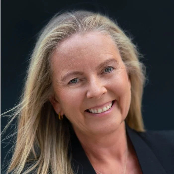 Close-up portrait of a smiling middle-aged woman with blonde hair and blue eyes, wearing a black blazer and small gold earrings, against a dark background.