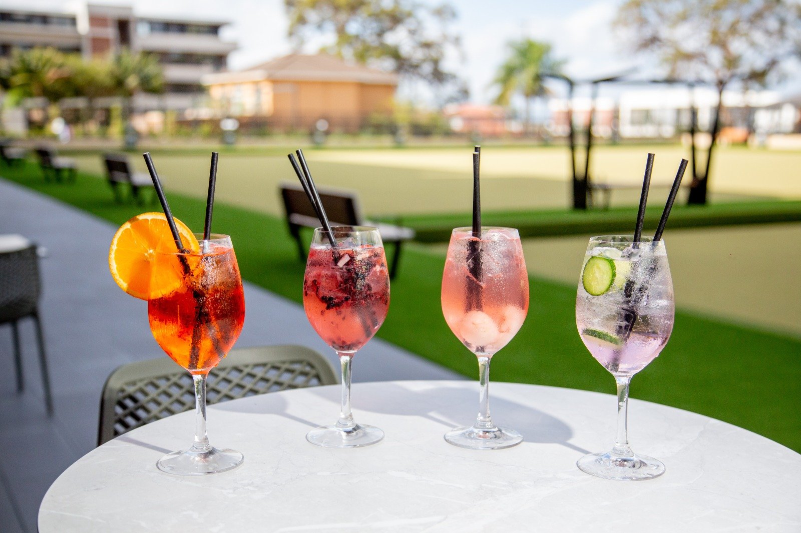 Four cocktails with ice and garnishes on a white table outdoors, with a grassy park, benches, and buildings in the background.