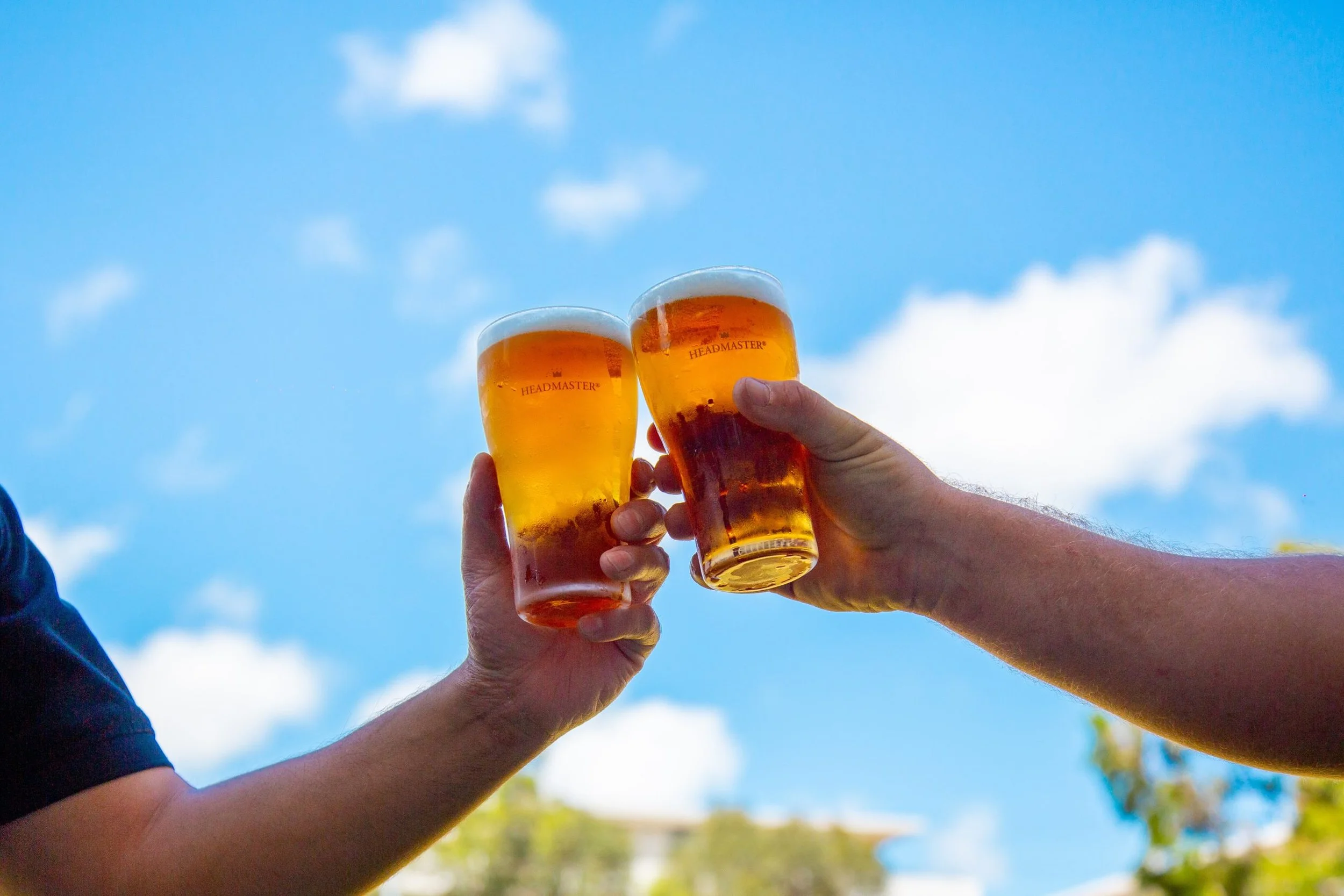 Multiple hands raising glasses of beer in a toast against a blue sky with clouds.
