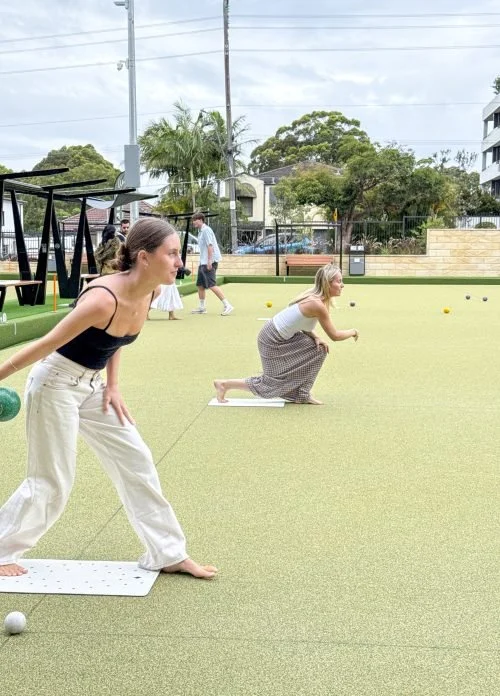 Two women practicing outdoor yoga on mats, one in a black tank top and white pants, the other in a white top and patterned skirt, with children in the background on a green outdoor space with trees and buildings.