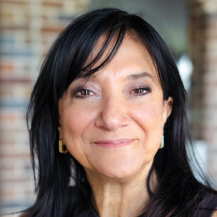 Close-up of a smiling middle-aged woman with black hair and earrings, standing outdoors with a blurred brick wall background.