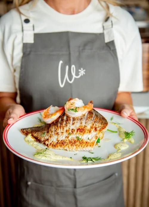 Person wearing a white shirt and a dark apron holding a plate with grilled fish topped with shrimp, garnished with herbs and sauces.