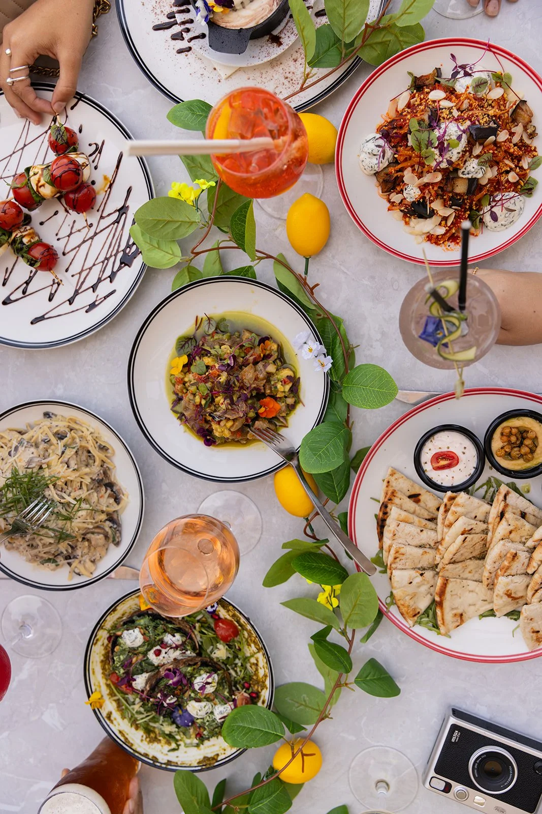 A top-down view of a table with various dishes, including a plate of cherry tomatoes with balsamic glaze and chocolate drizzle, a bowl of pasta, a salad with vegetables and flowers, and a plate of grilled flatbread with dips. There are also drinks with straws, a camera, and yellow lemons on the table decorated with green leaves and small yellow flowers.