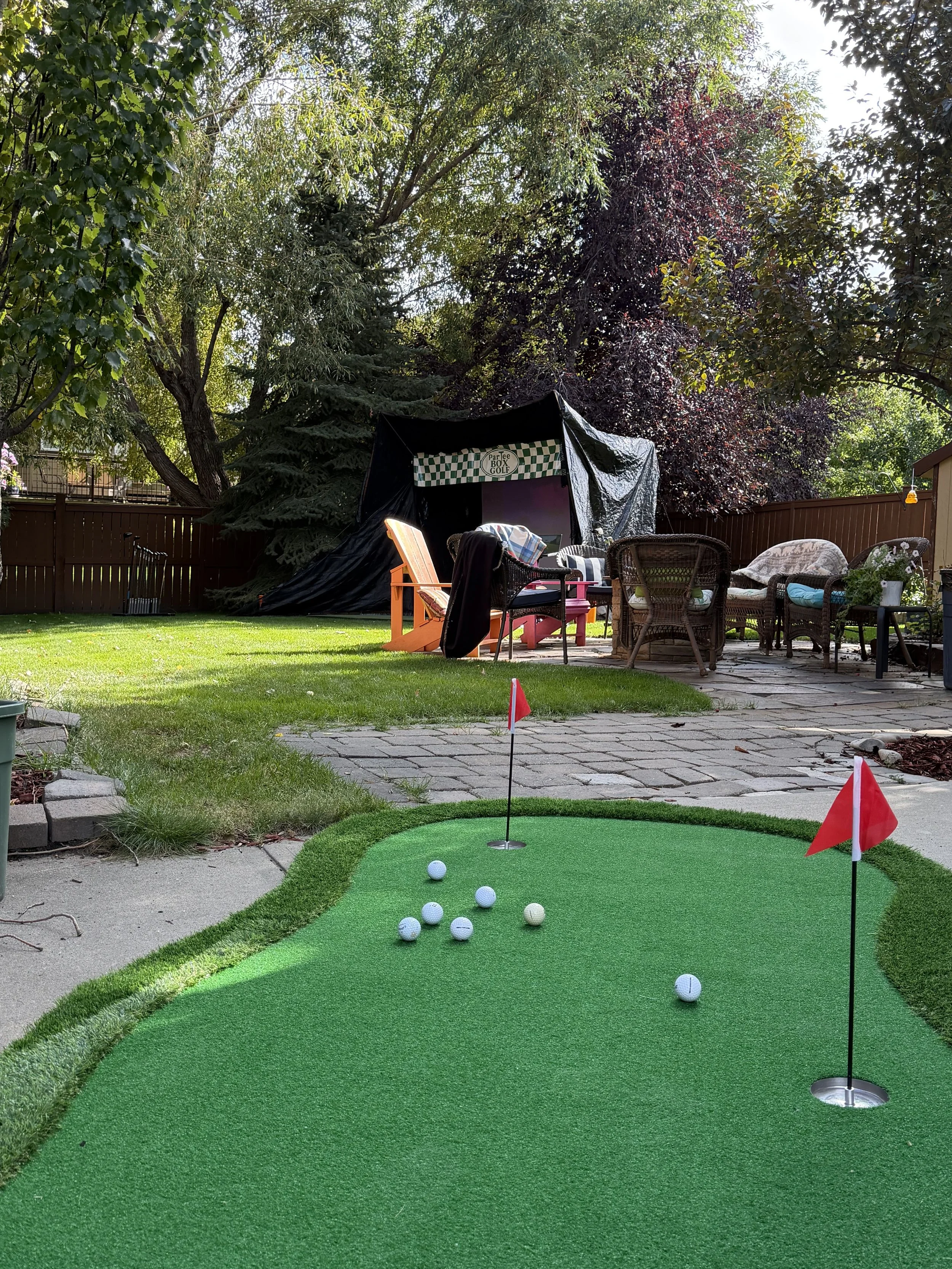 A backyard with an outdoor seating area, a mini golf putting green with seven golf balls and two red flags, surrounded by trees and a wooden fence.