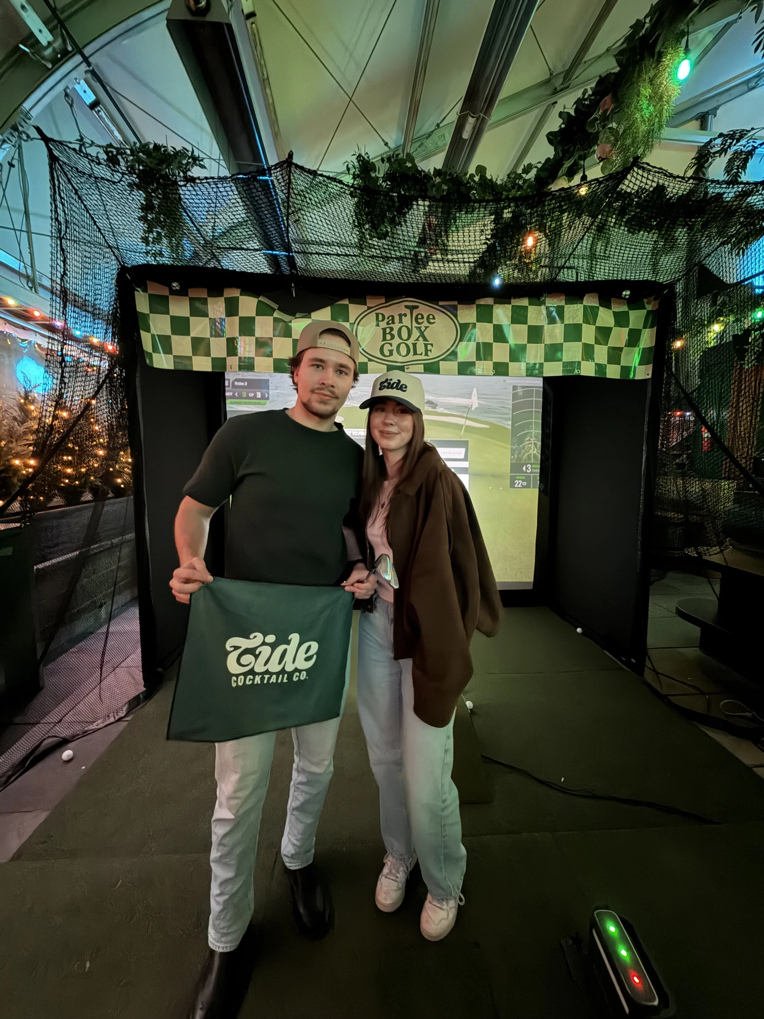 Two people standing in front of a golf simulation screen at the ParTee Box Golf indoor venue, holding a green bag with the Tide Cocktail Co. logo. The man on the left is wearing a gray cap, black t-shirt, and light-colored pants. The woman on the rig
