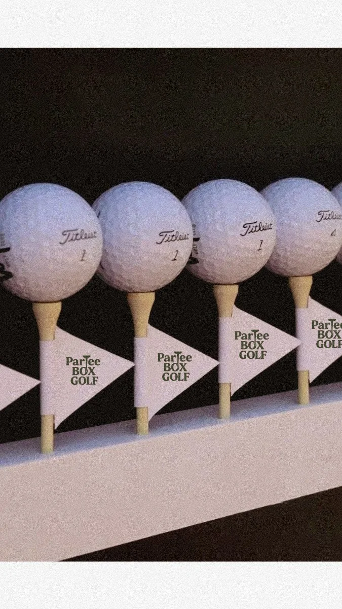 Golf balls resting on top of tee markers with 'ParTee Box Golf' written on them, arranged in a row on a golf course.
