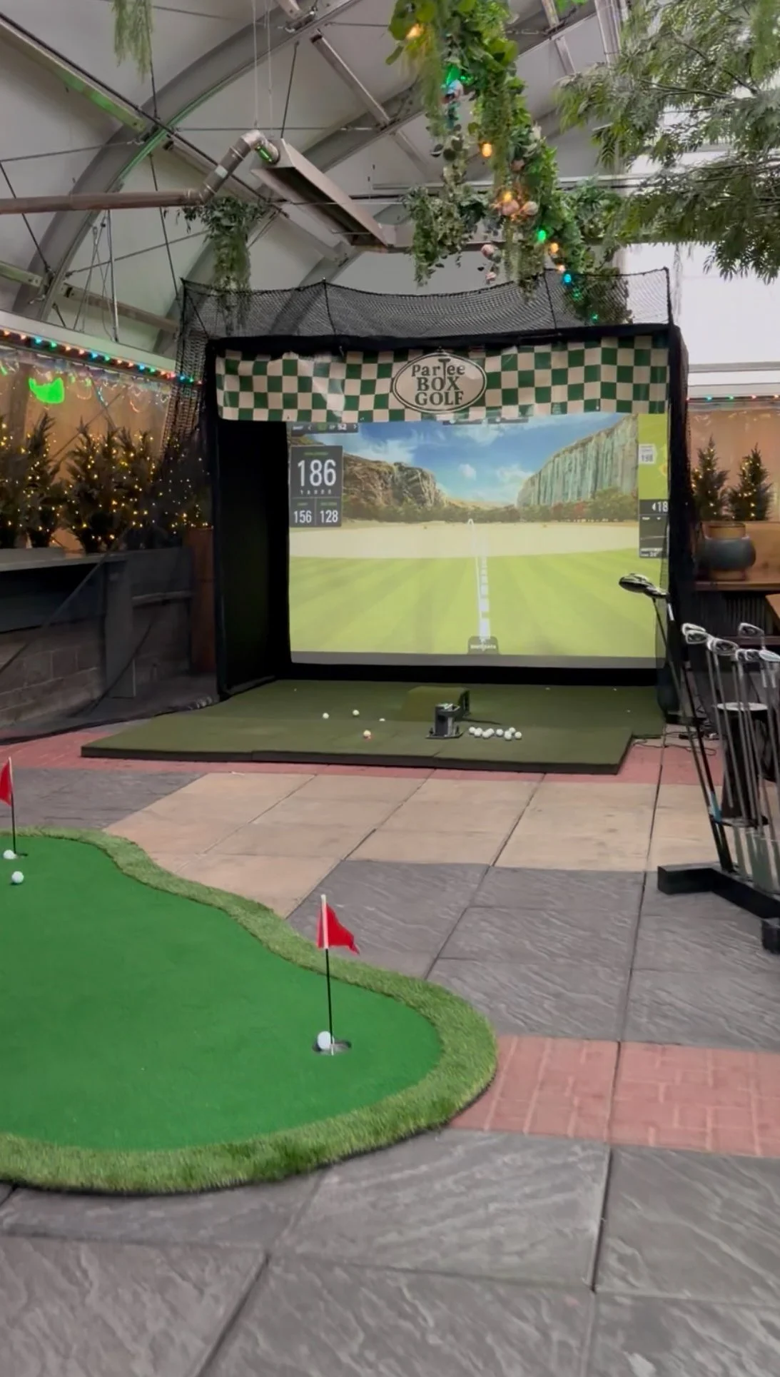 Indoor golf simulator setup with a screen displaying a golf course, a small putting green with flags and golf balls, and various golf clubs on the right side.