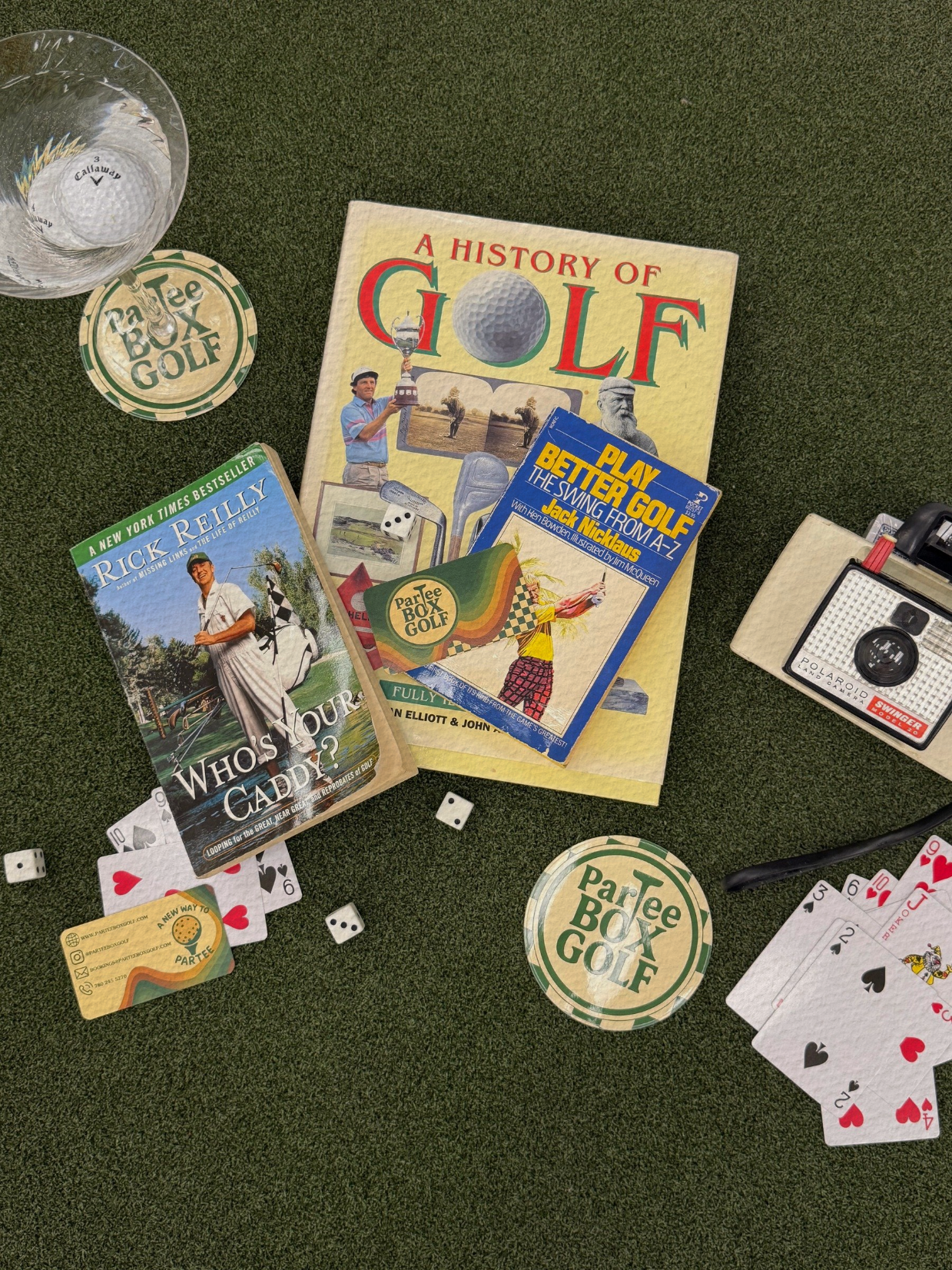 Collection of golf-themed books, playing cards, billiard balls, a glass of water, and a Polaroid camera on green artificial turf.