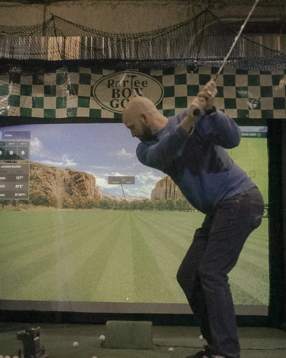Man practicing golf swing indoors in front of a virtual golf simulator screen with a scenic outdoor golf course background.