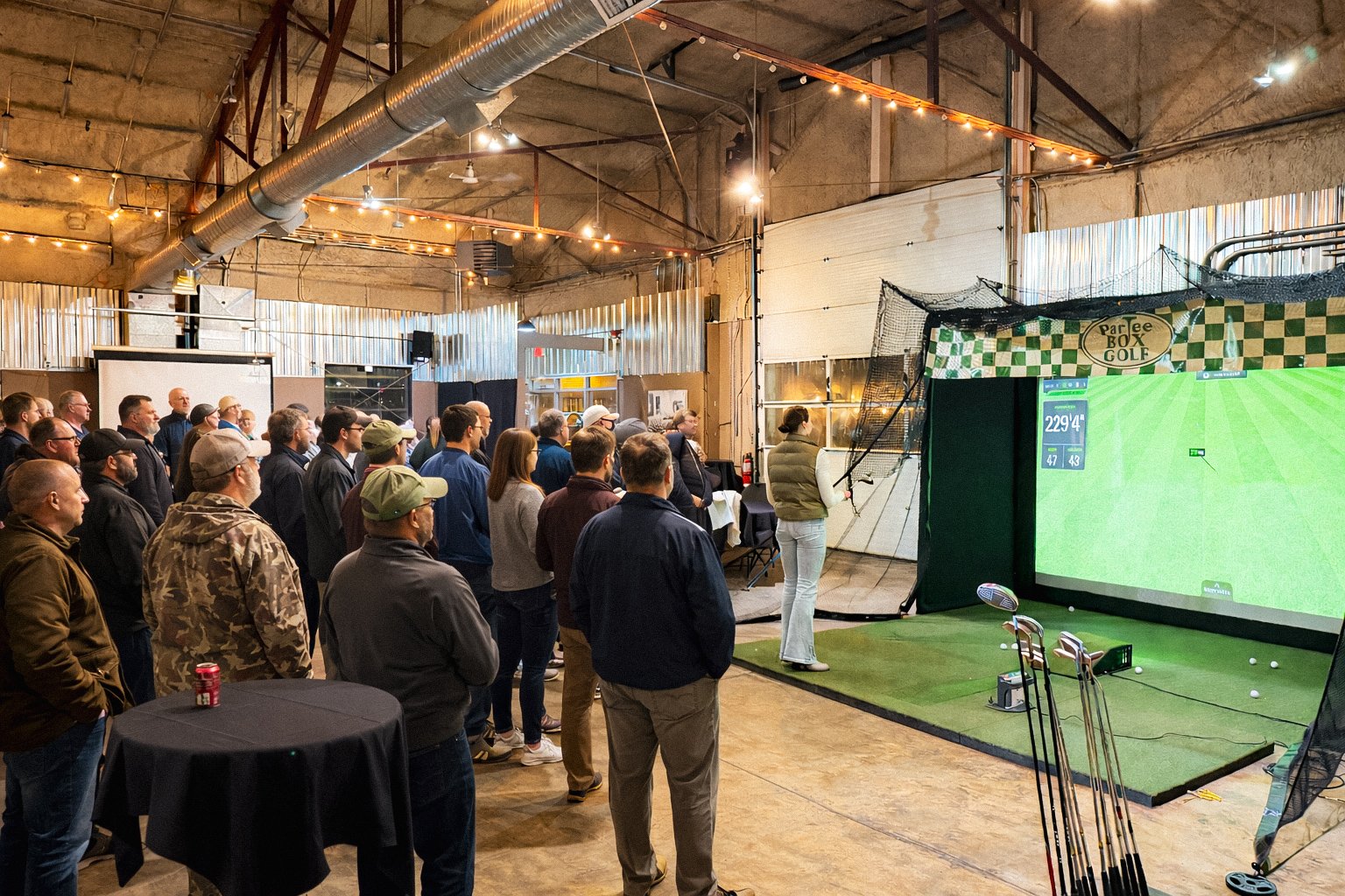 A group of people standing and watching a golf simulator inside a large, rustic indoor venue with string lights and metal walls. A woman in a vest is hitting golf balls at the simulator screen.