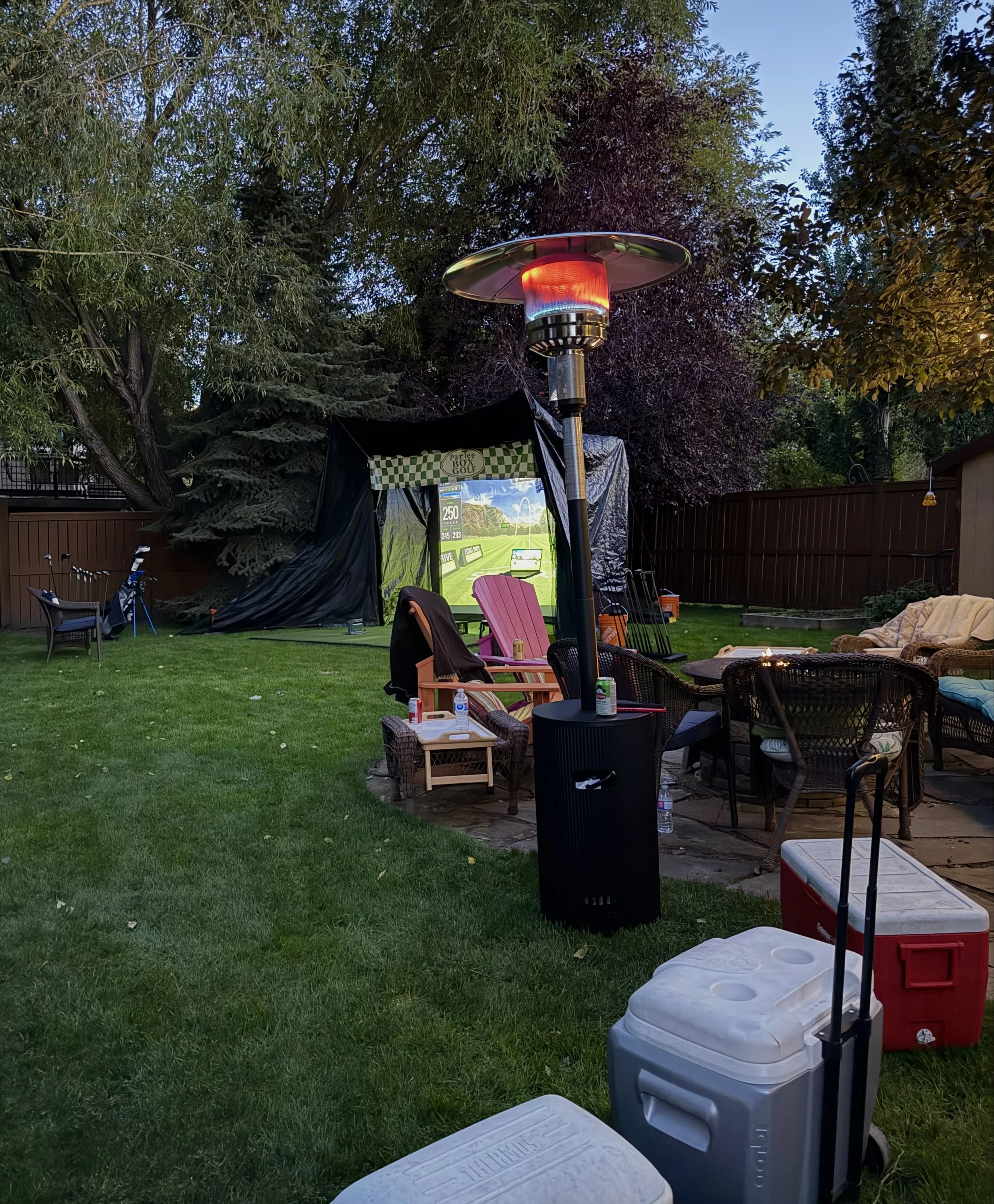 Backyard with outdoor furniture, a large patio heater emitting heat, a screen showing a golf game, and various coolers and chairs surrounded by trees at dusk.