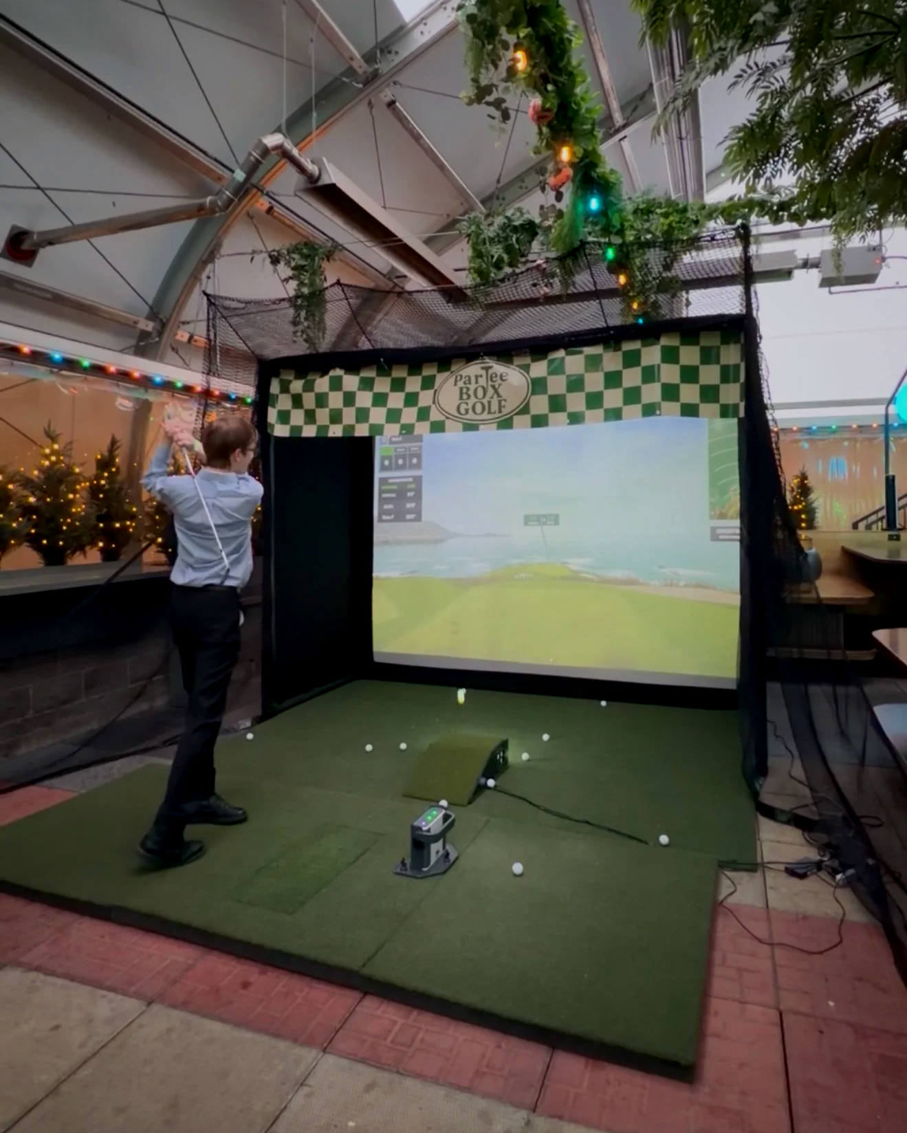 A person practicing golf swings indoors on a simulated golf screen at a golf simulation setup with artificial turf, golf balls on the ground, and a large screen displaying a golf course scene.