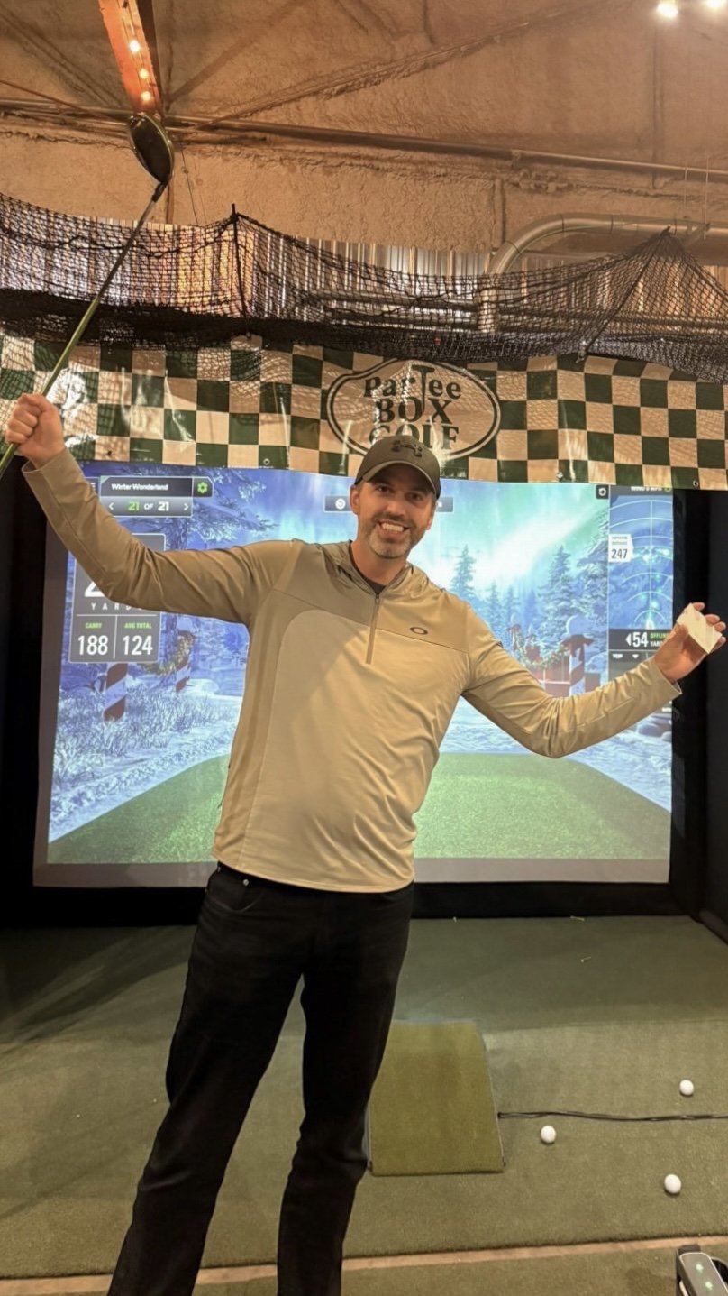 A man in a beige long-sleeve shirt, black pants, and a cap is at the indoor golf simulation, holding a golf club in one hand and a scorecard in the other, standing on a small grassy mat with golf balls nearby, smiling at the camera.
