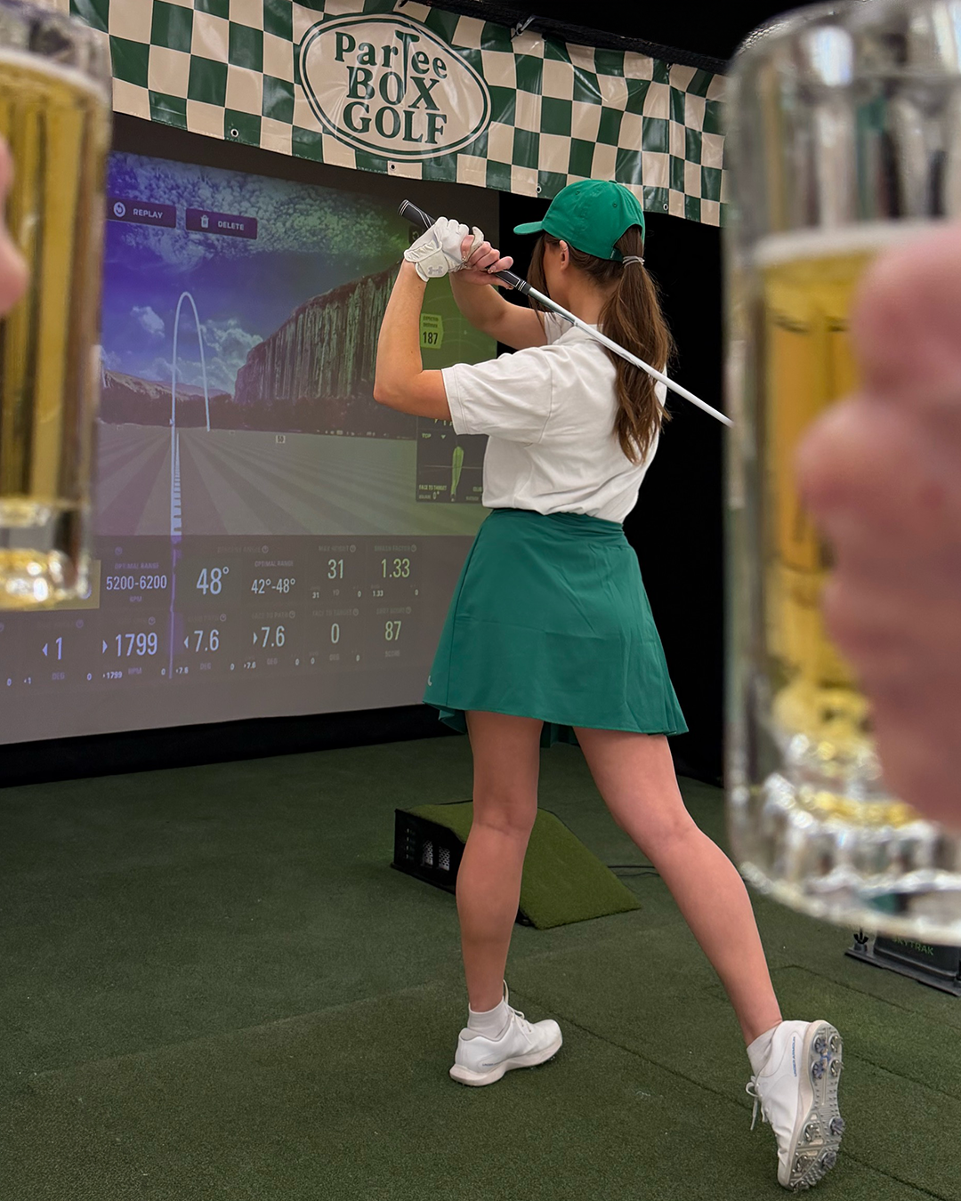 A woman in golf attire, including a green cap, white shirt, green skirt, and golf shoes, is playing virtual golf on a simulator. She is in a golf stance, swinging a golf club with a large screen displaying a virtual golf course in front of her. The background has a sign that reads "Par Tee Box Golf," and the scene is set indoors with artificial turf and bottles of beer in the foreground.