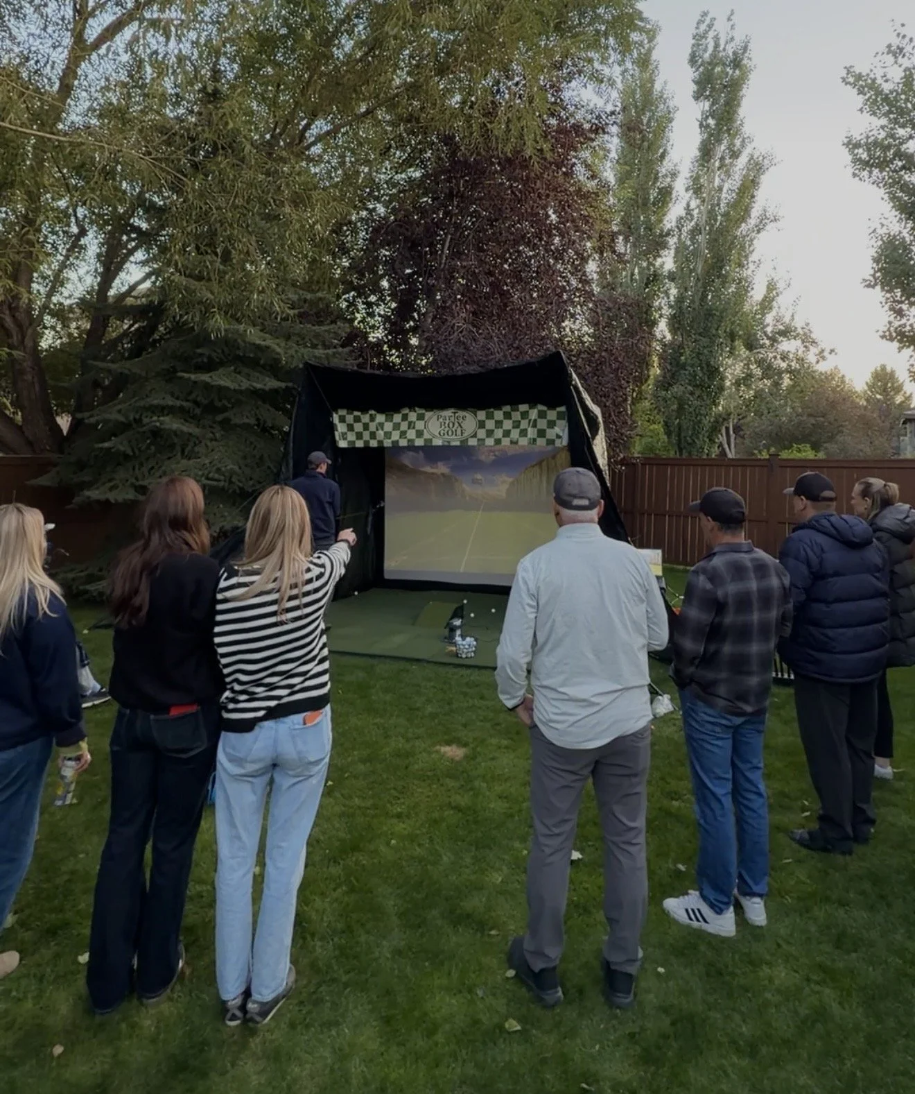 People gathered outdoors watching a golf simulation on a large screen, with trees and a wooden fence in the background.