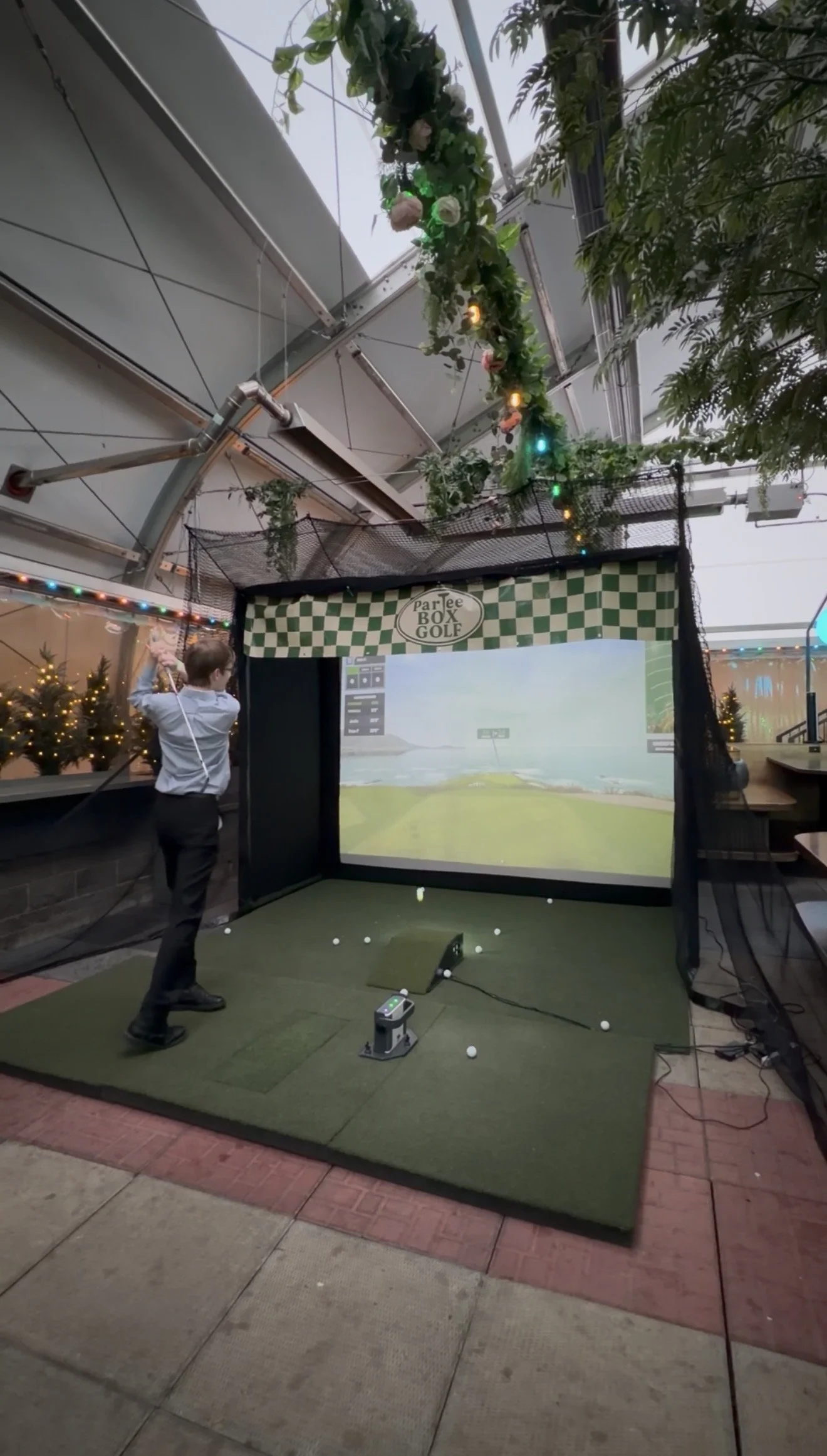An indoor golf simulator setup with a young boy practicing his swing. The simulator has a screen displaying a virtual golf course, surrounded by a protective net and decorated with string lights and plants.