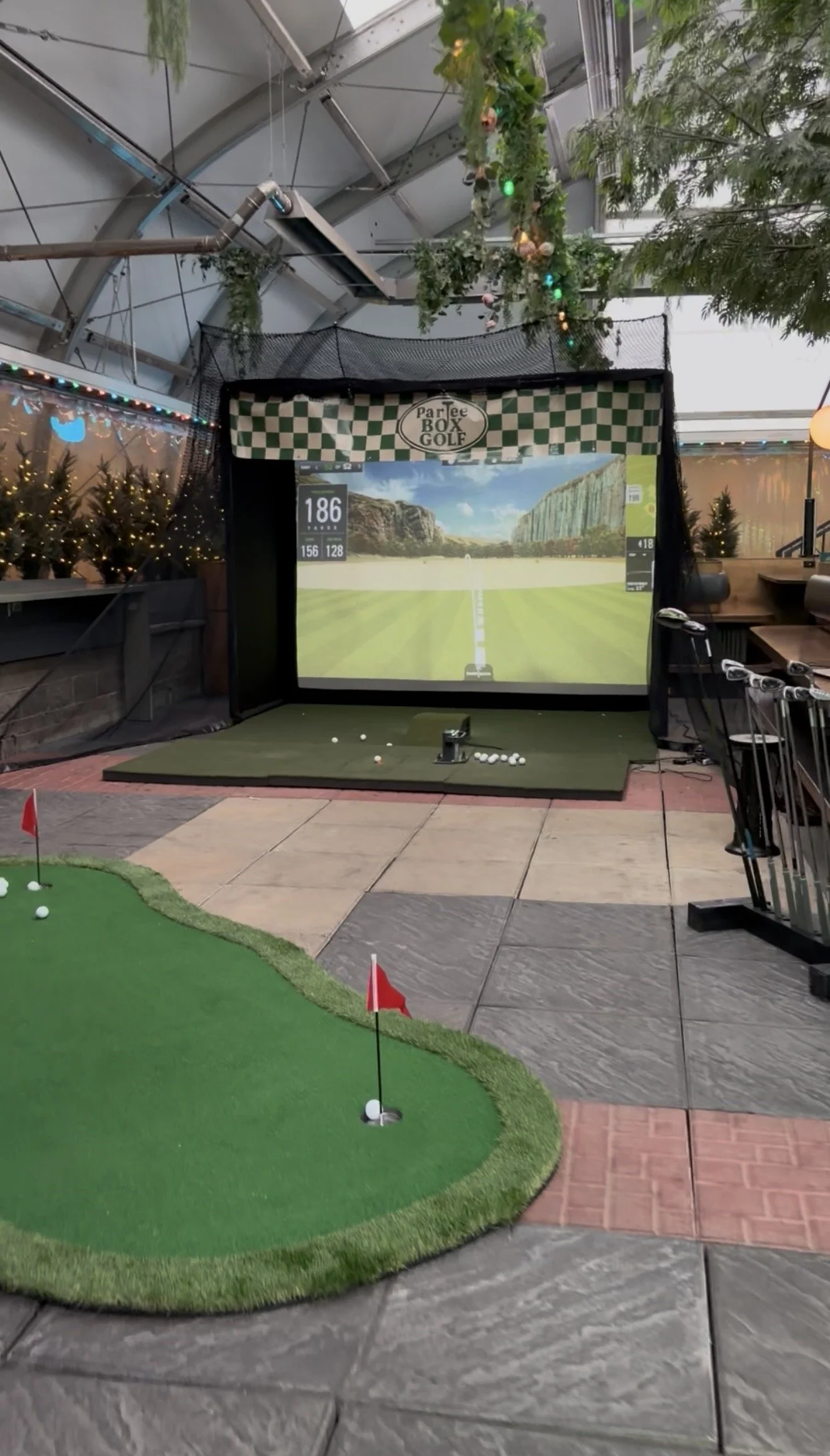 Indoor golf simulation setup with a large screen showing a golf course, a putting green with flags and golf balls, and golf clubs on the right side.