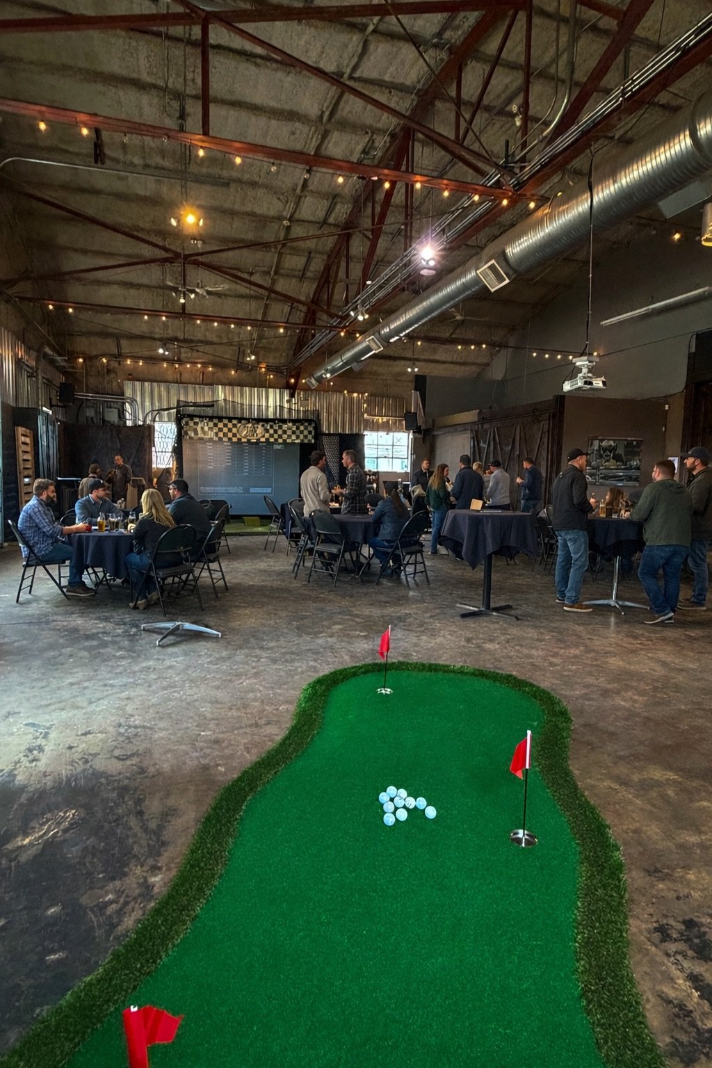 An indoor bar or event space with a mini golf putting green in the foreground, and groups of people socializing at tables, around tall tables, and standing in line at a bar area. The ceiling has industrial elements like exposed ductwork and string lights.
