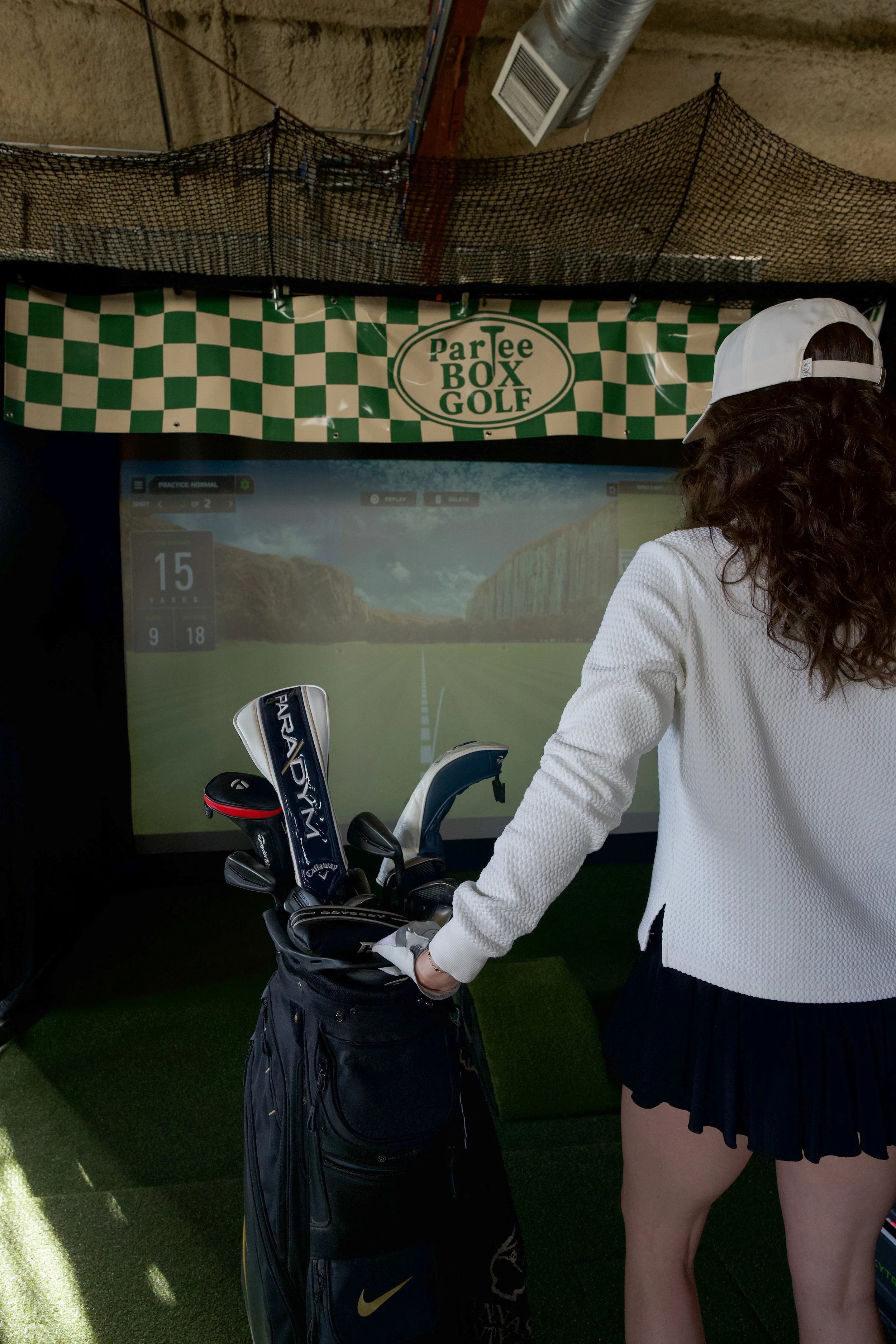 Woman standing in front of a golf simulator screen with a golf bag containing clubs. The simulator screen displays a virtual golf course scene.