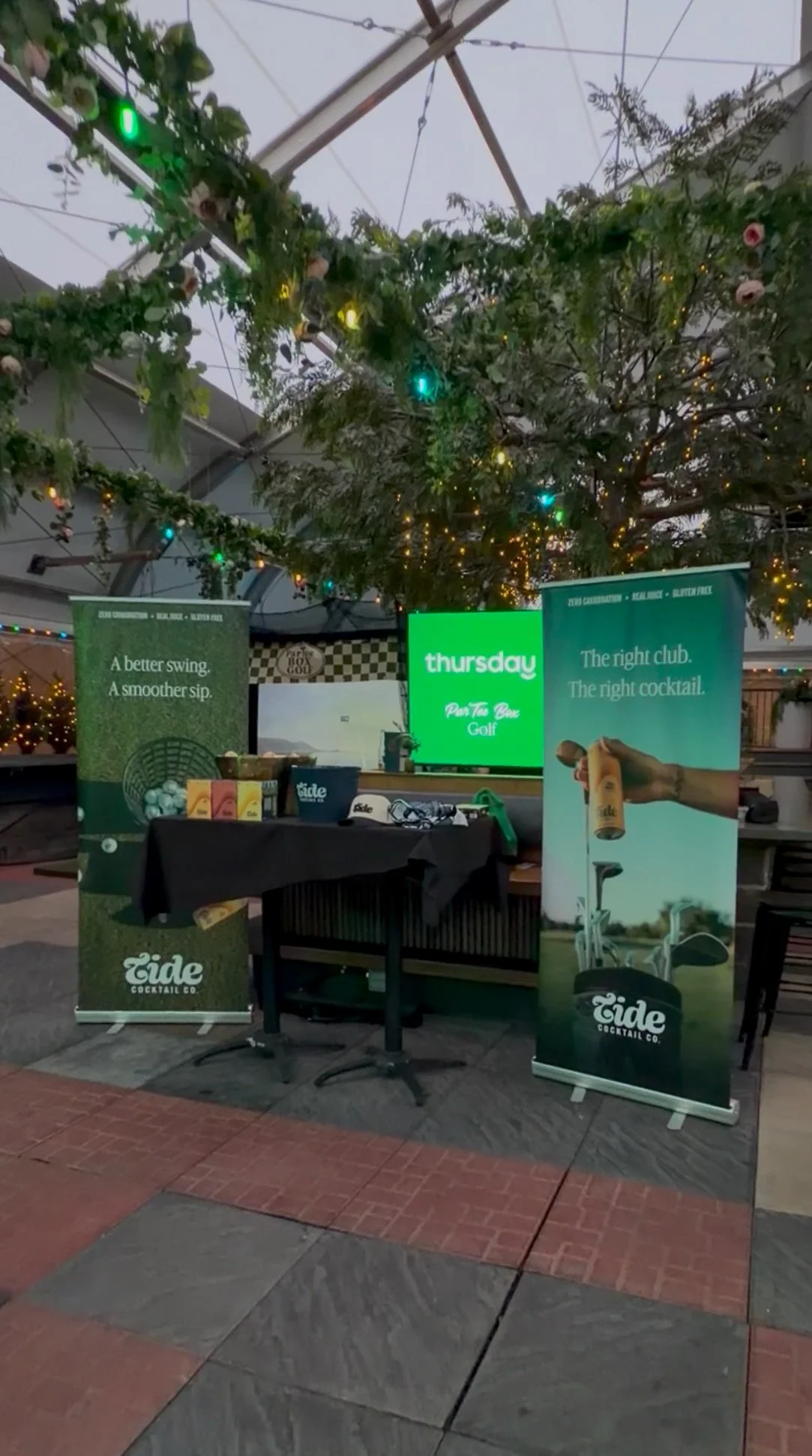 An indoor setup with a black table covered with various promotional items and banners in front of a green screen displaying 'Thursday'. The banners are advertising Eide Cocktail Co with slogans about golf and cocktails. The ceiling has string lights 