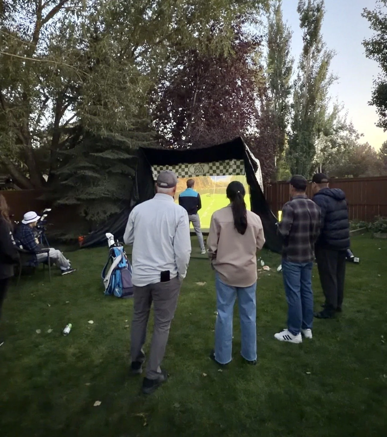 People watching a golf simulacrum outdoors in a backyard.