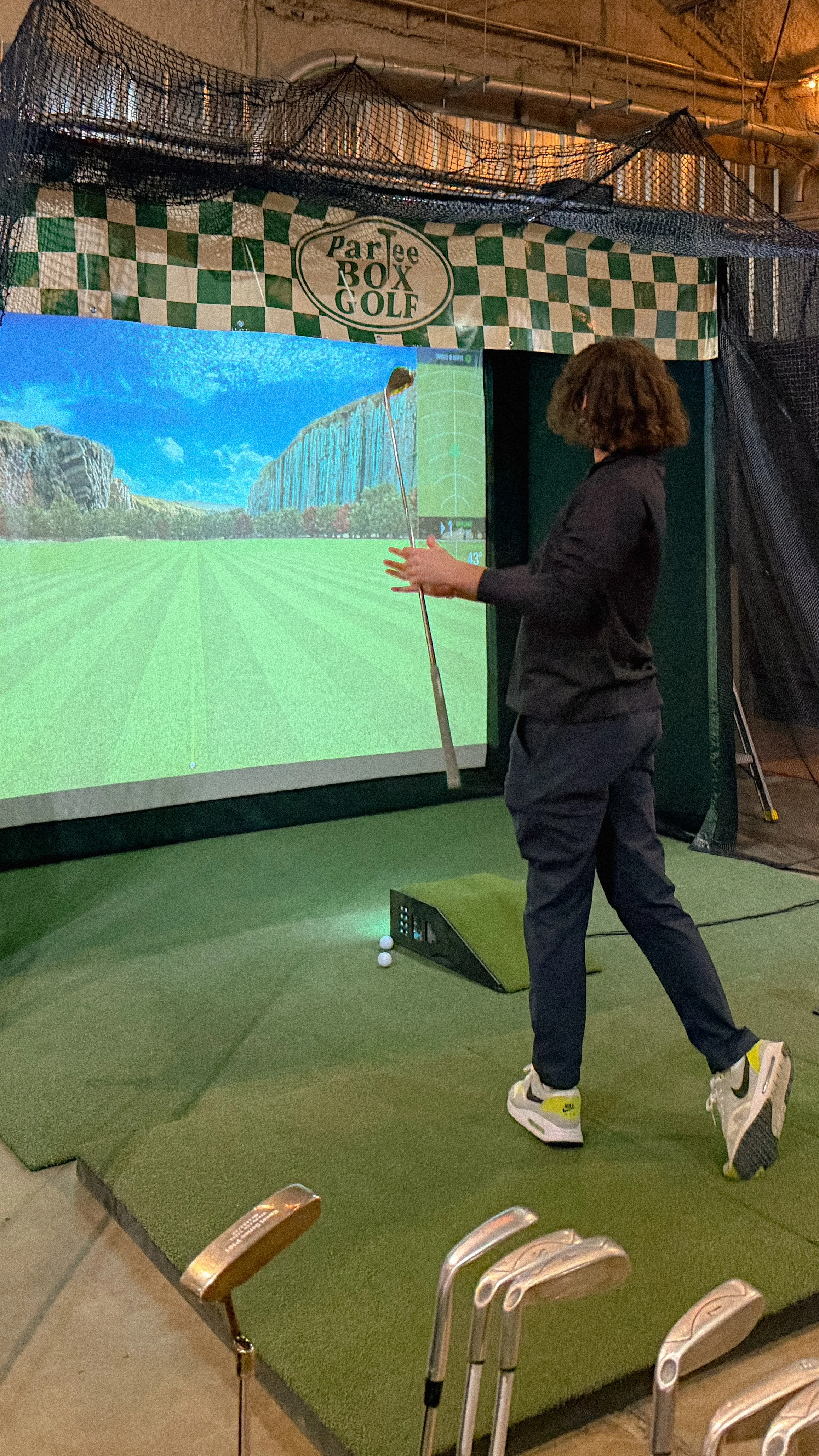 Woman playing virtual golf at indoor golf simulator with a large screen showing a golf course.