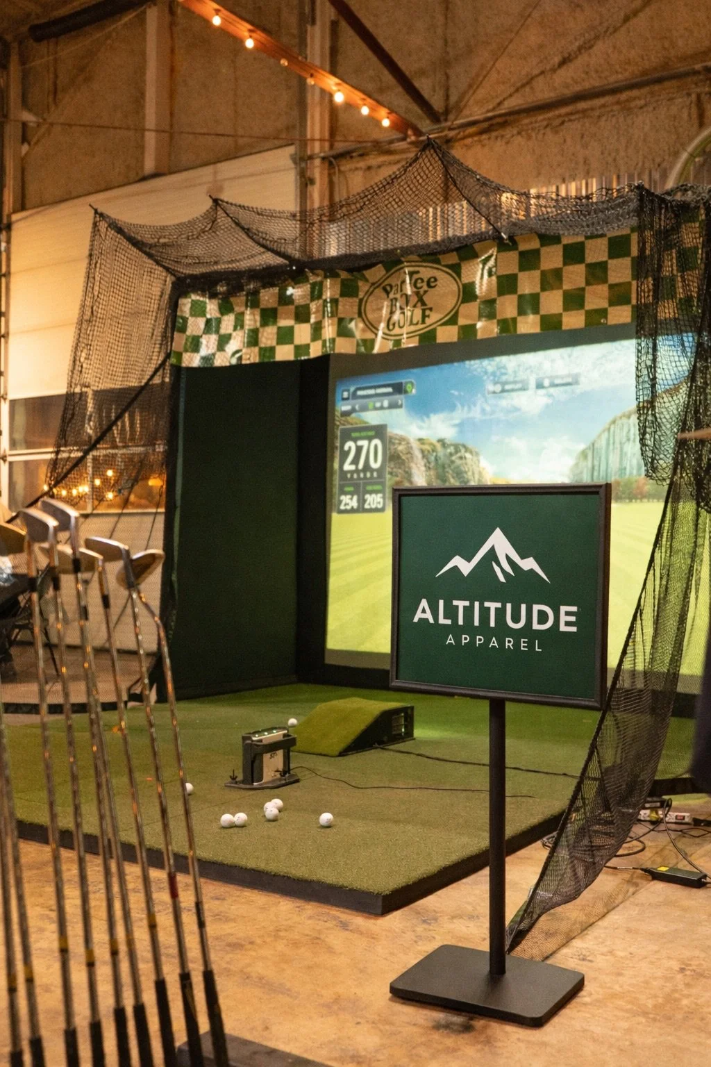 Indoor golf simulator setup with a screen displaying a golf course, a sign for "Altitude Apparel," and golf balls on a simulated turf mat surrounded by safety nets.