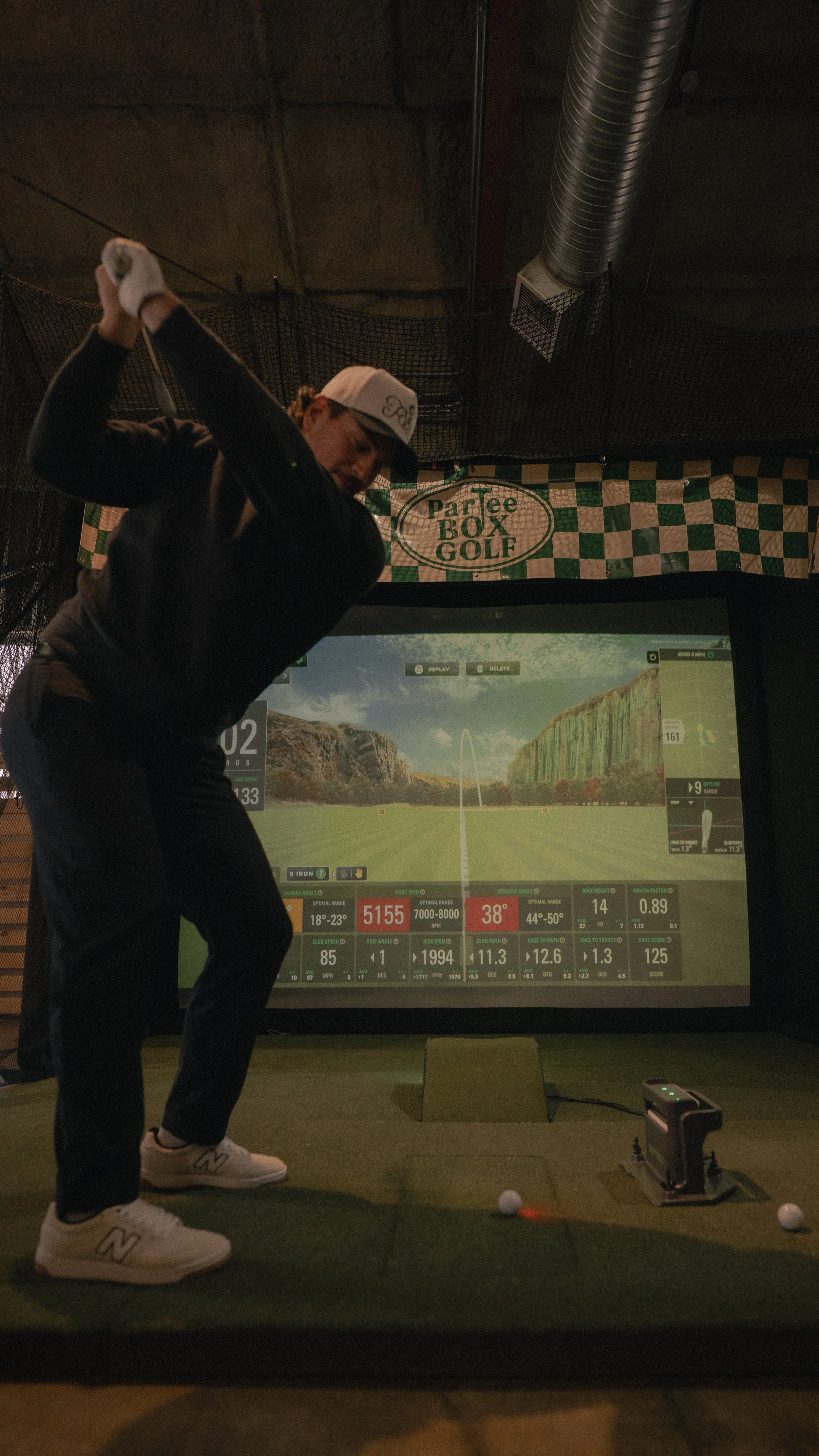 Person practicing golf swings in an indoor golf simulator with a large screen displaying golf course data.