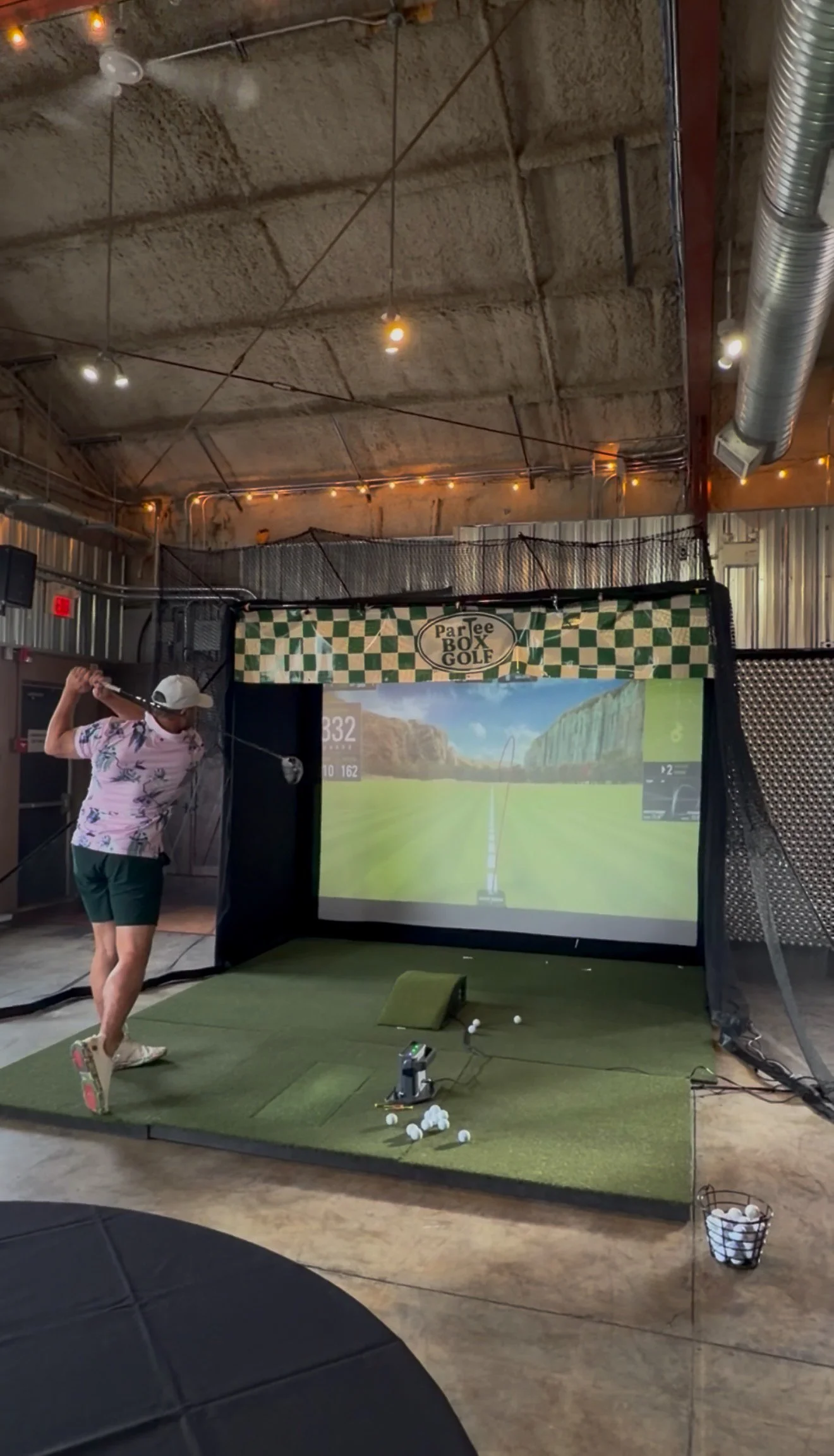 An indoor golf simulator setup with a person practicing a swing, multiple golf balls on the floor, a large screen displaying a virtual golf course, and signs reading 'ParTee Box Golf'.