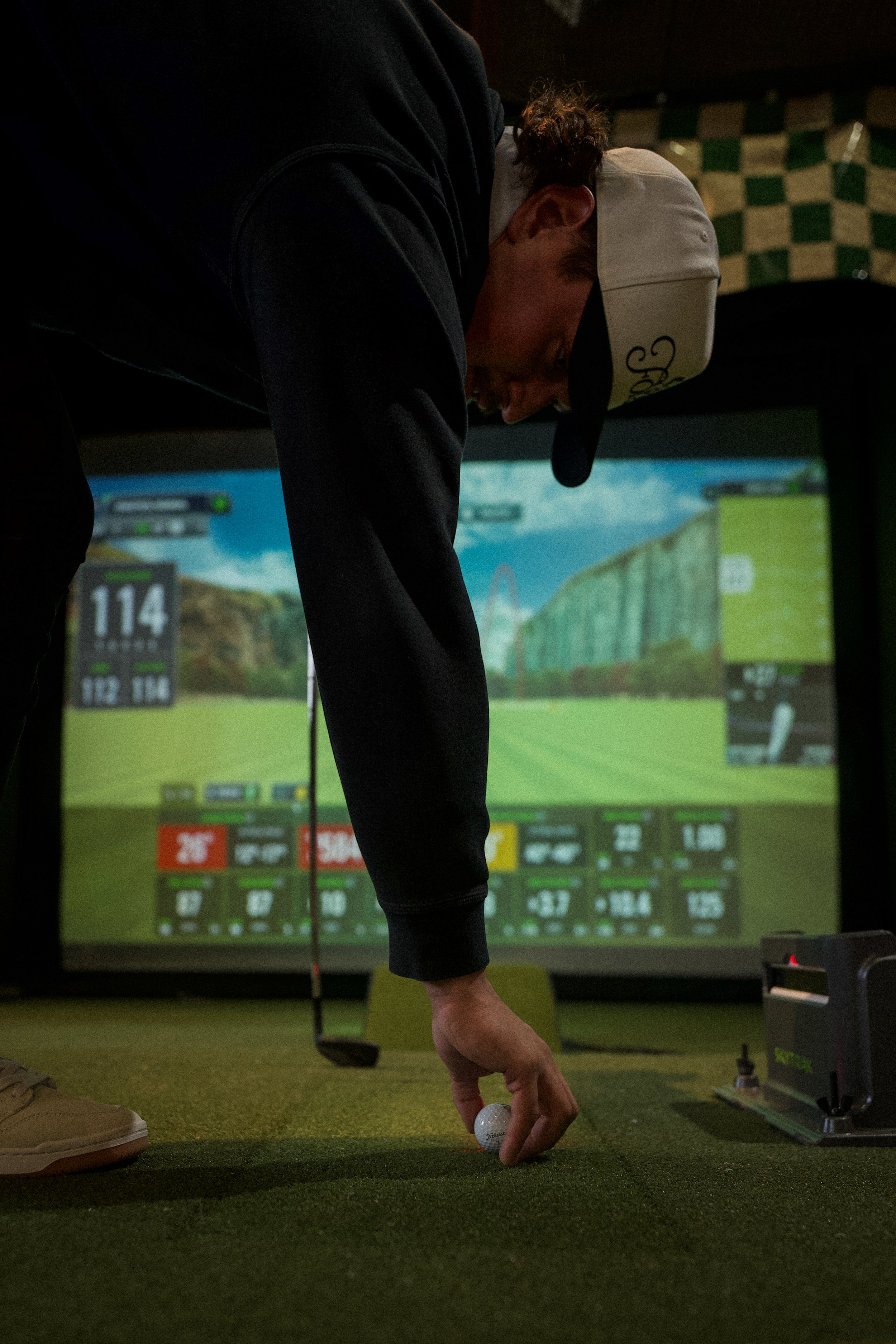 Person in a black hoodie and beige cap preparing to hit a golf ball in an indoor golf simulation room.