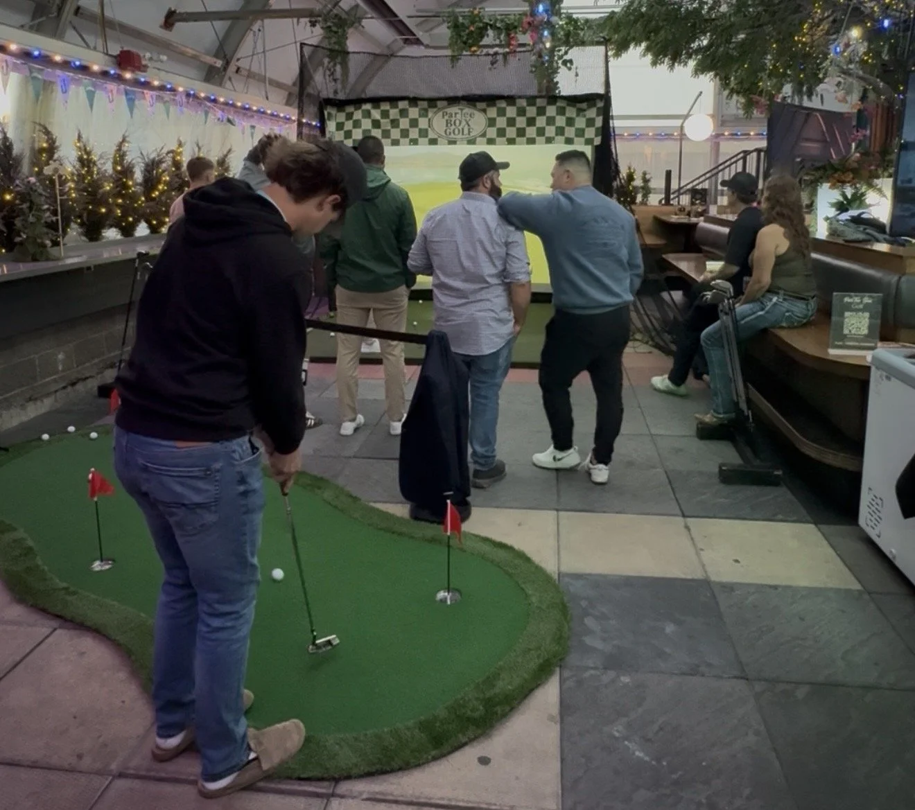 People playing mini golf indoors, with a man preparing to putt and others watching, festive decorations and string lights in the background.