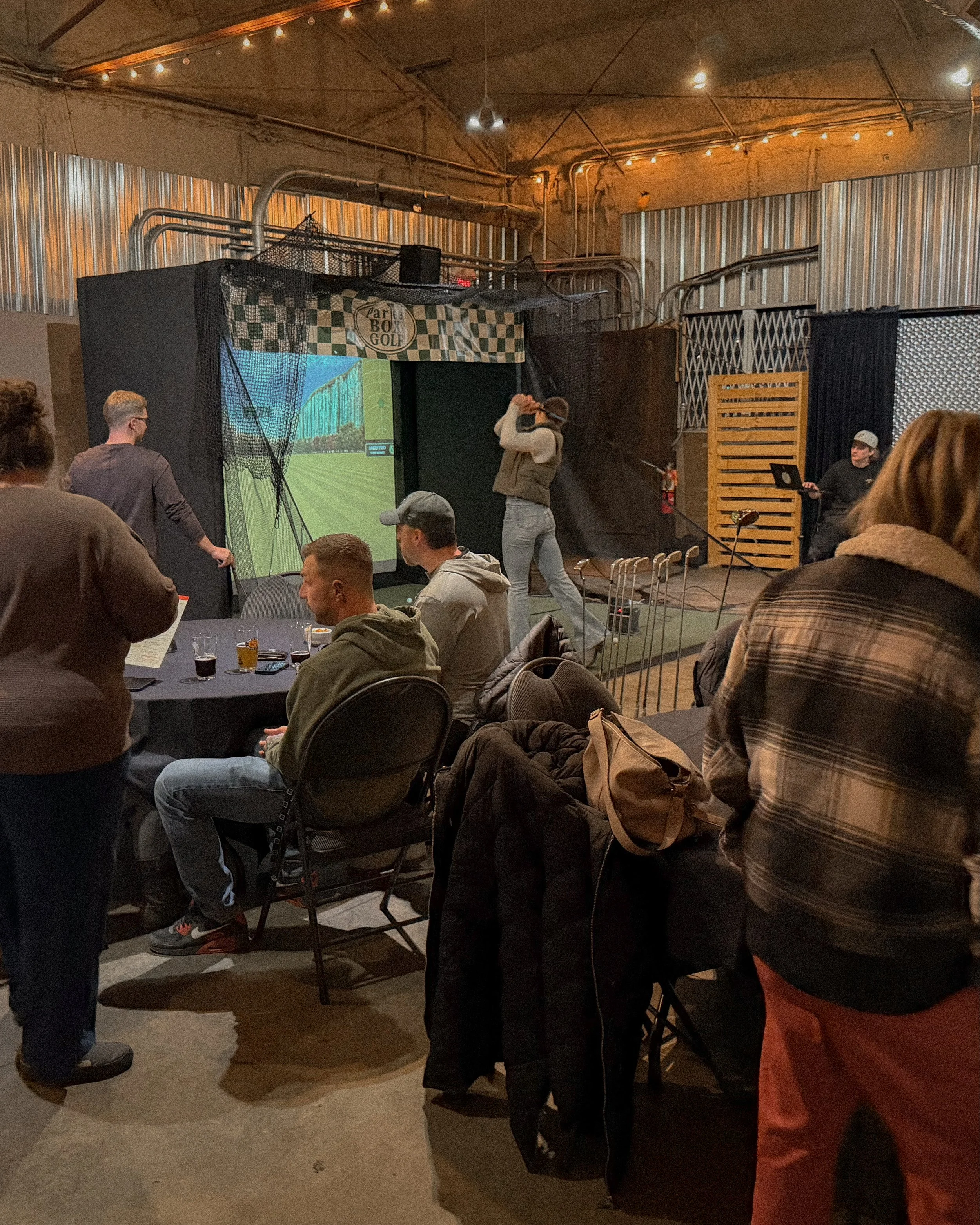 People playing a virtual golf game in an indoor arcade, with a woman swinging a golf club and others seated around tables.
